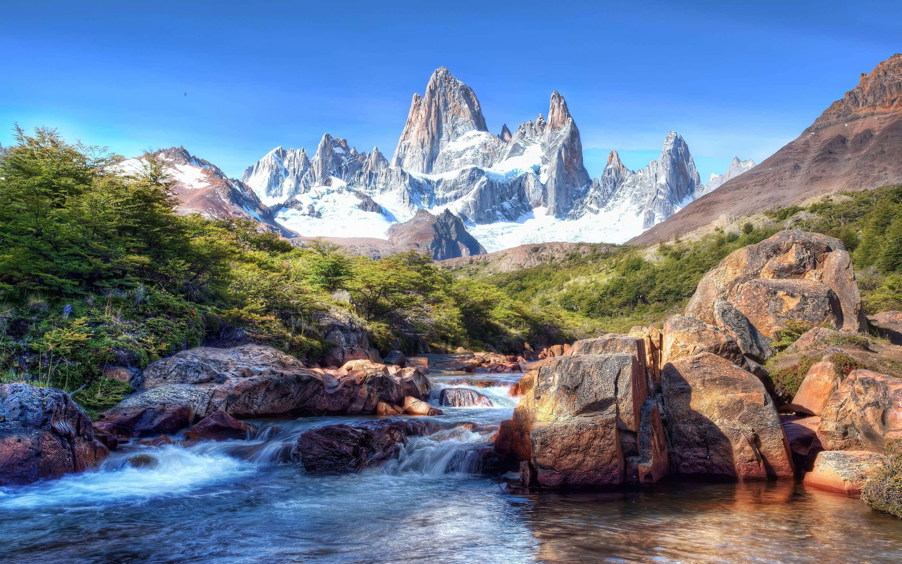 Patagonia 桌布 壁紙 — 一條湍急的河流在崎嶇的岩石上流過，下方是清澈的藍天，上方是高聳的雪山峰頂。