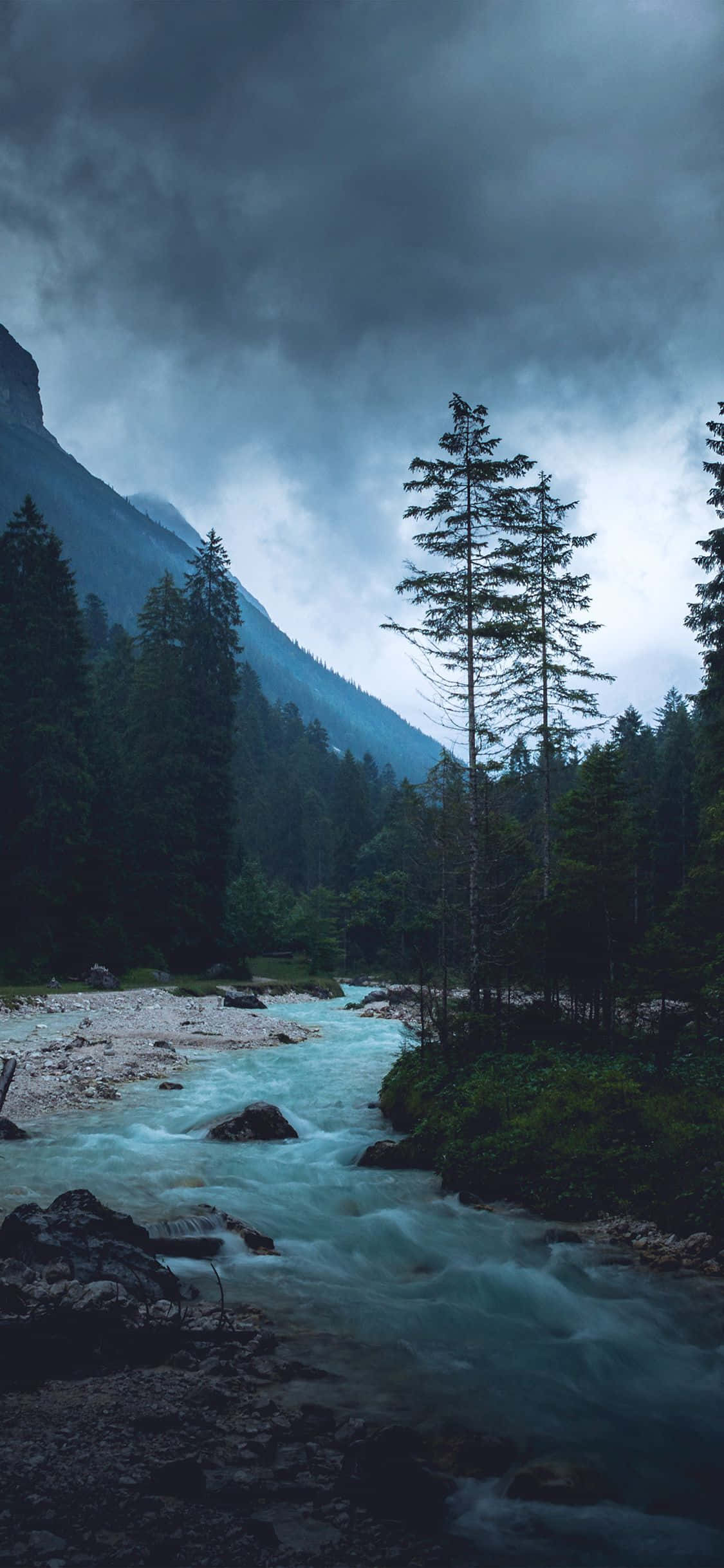 iPhone X Nature 桌布 壁紙 — 一條湍急的河流在暴風雨灰暗的天空下，穿過黑暗的森林山谷，背景是高聳的山脈。
