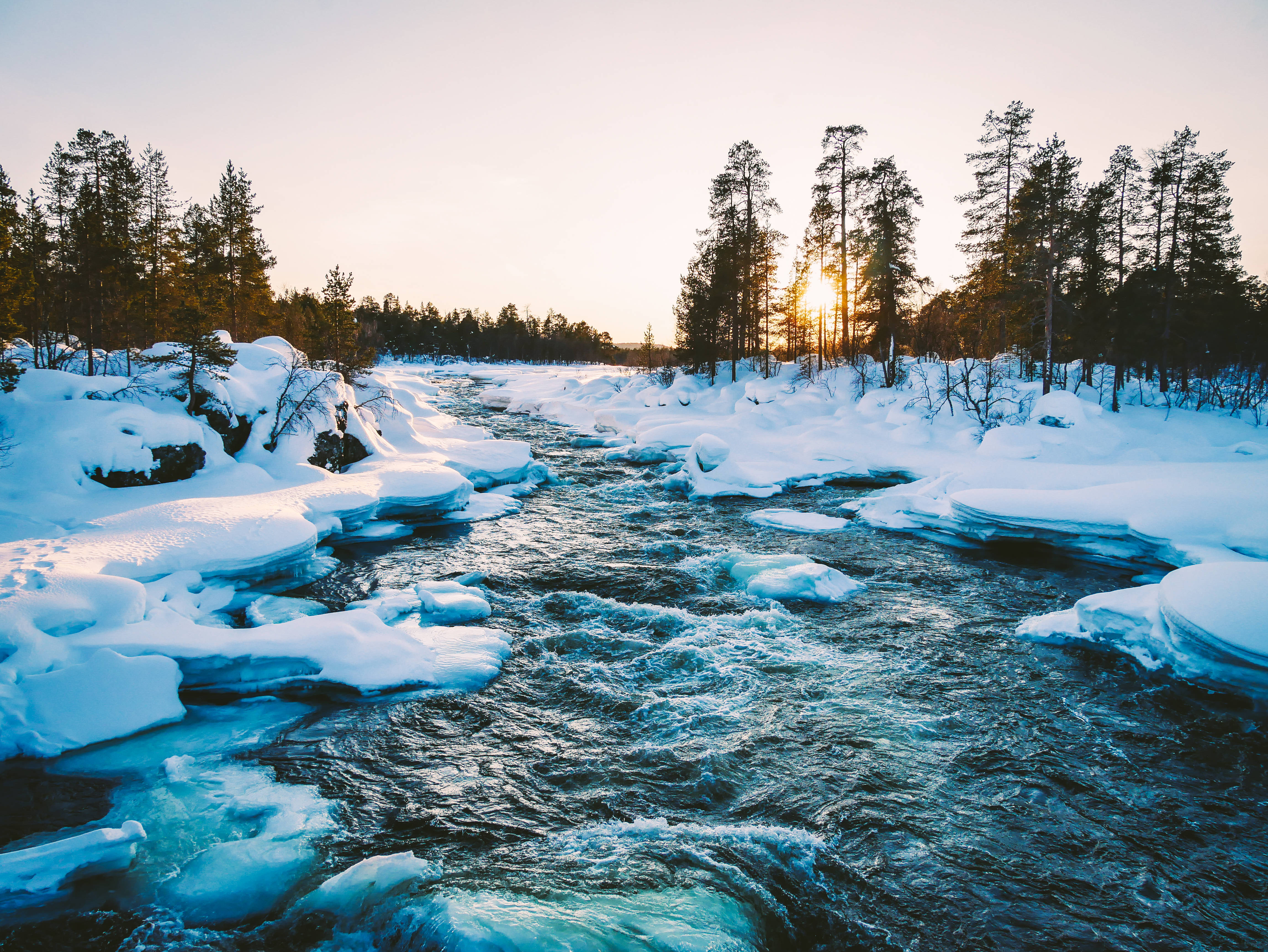 Finland 桌布 壁紙 — 一條湍急的河流穿過白雪覆蓋的冬季森林，夕陽落在高大的松樹後面，灑下金色的光芒。