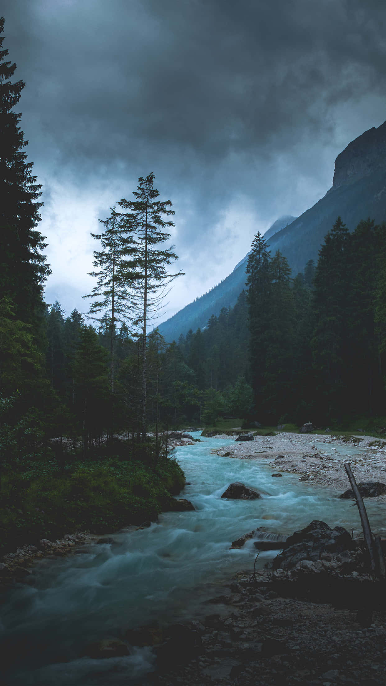 黑暗自然 桌布 壁紙 — 一條湍急的河流穿過黑暗的森林山谷，背景是暴風雨灰濛濛的天空和巍峨的山脈。