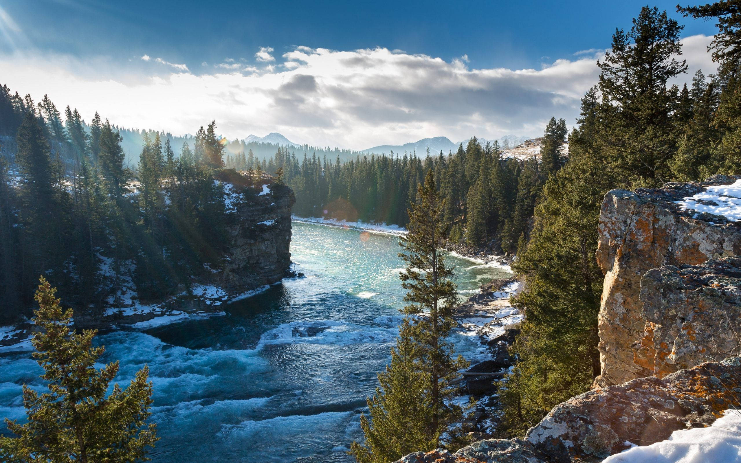山景 桌布 壁紙 — 一條湍急的碧綠河流穿過白雪覆蓋的懸崖和常綠森林，在明亮的藍天和散落的雲朵下。