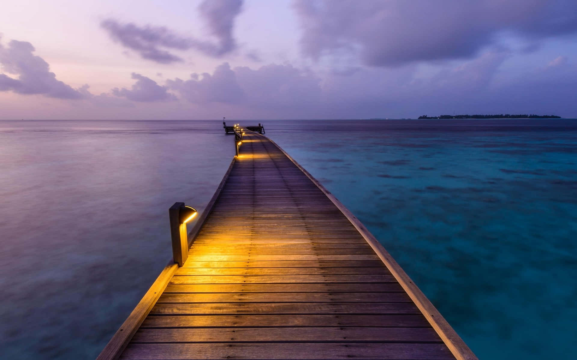 Calming Desktop 桌布 壁紙 — 一條溫暖照明的木製碼頭延伸至藍綠色海水，背景是紫色黃昏天空，散落著雲朵。