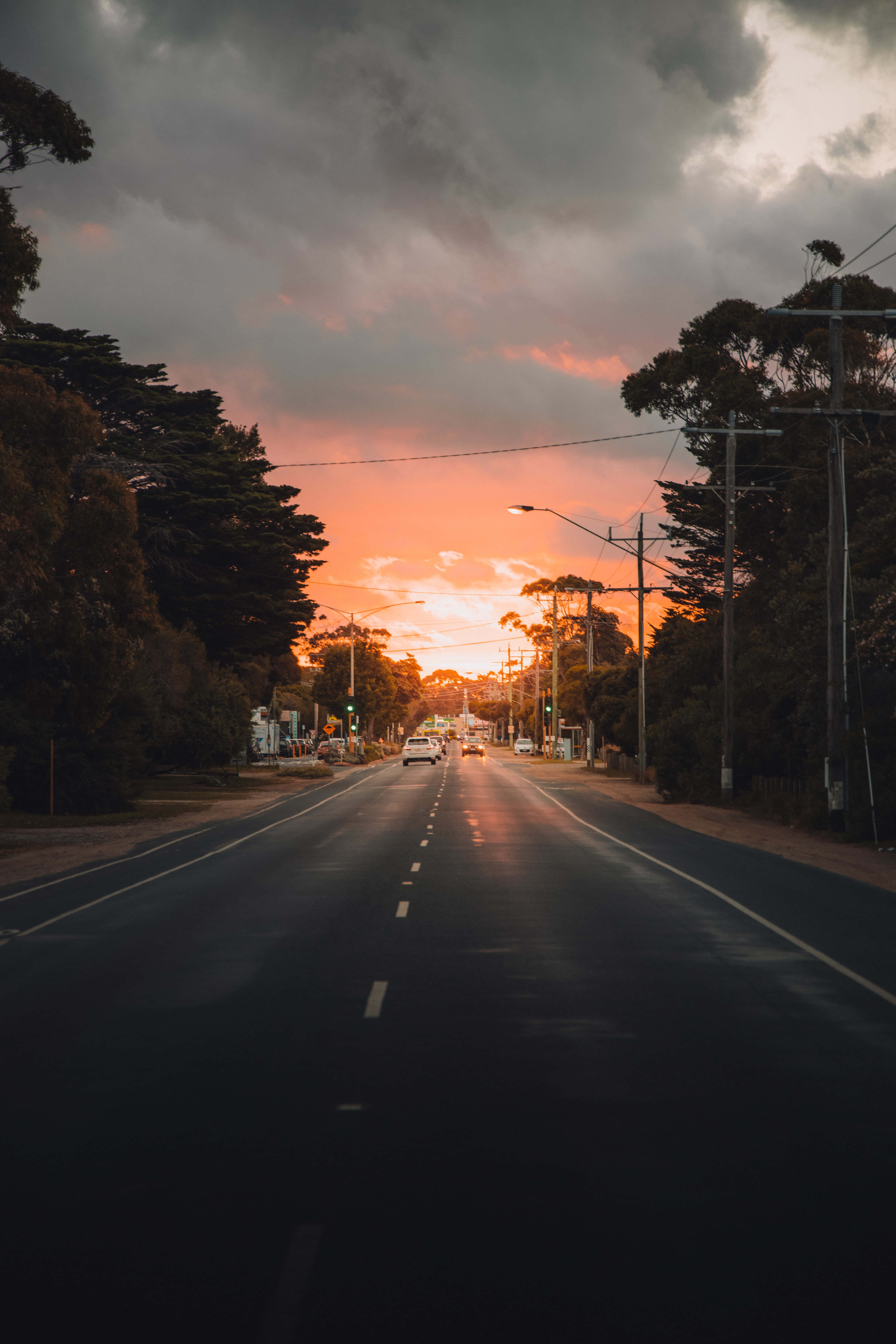 Street 桌布 壁紙 — 一條濕漉漉的道路伸向一個充滿活力的橙色日落，兩旁是黑暗的樹木和電線桿，上方是戲劇性的雲層。