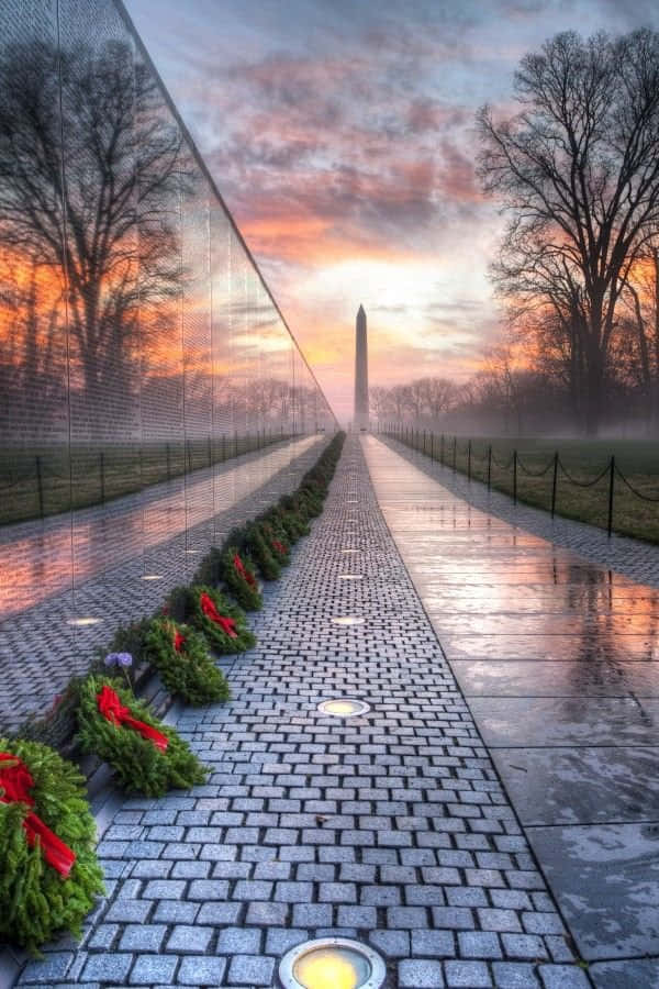 Memorial 桌布 壁紙 — 一條濕漉漉的鵝卵石小徑兩旁擺放著花圈，在黎明時分通往華盛頓紀念碑，倒映在越戰紀念碑牆上，天空呈現柔和色調。