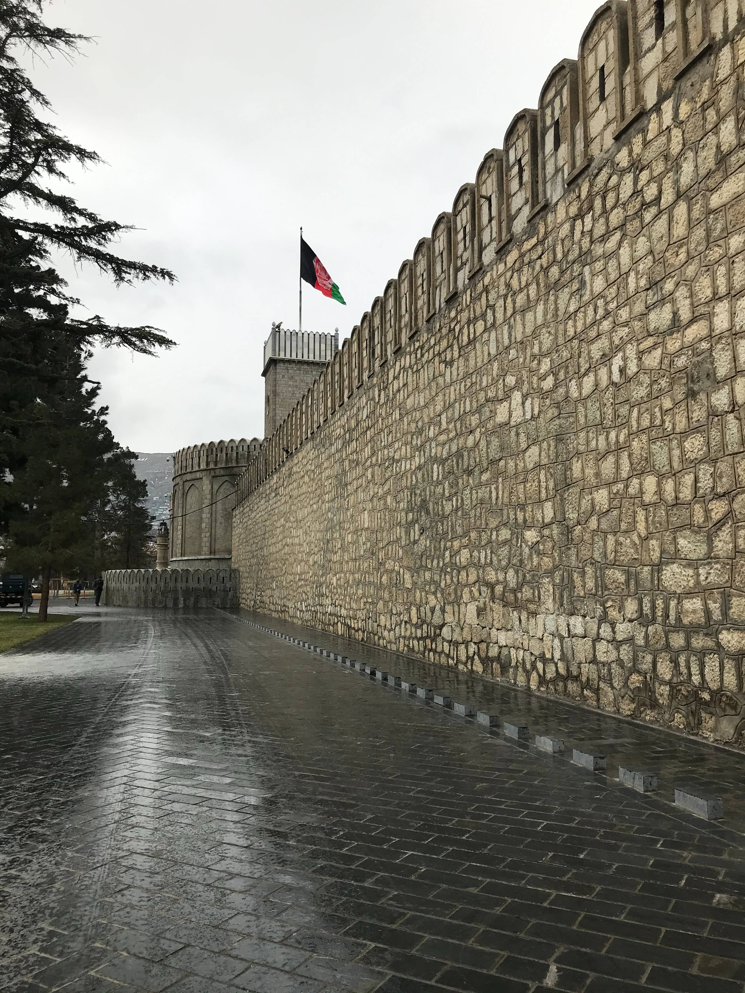 Kabul 桌布 壁紙 — 一條濕漉漉的鵝卵石小路，沿著高聳的古老石頭堡壘城牆延伸，天空多雲，塔頂飄揚著旗幟。