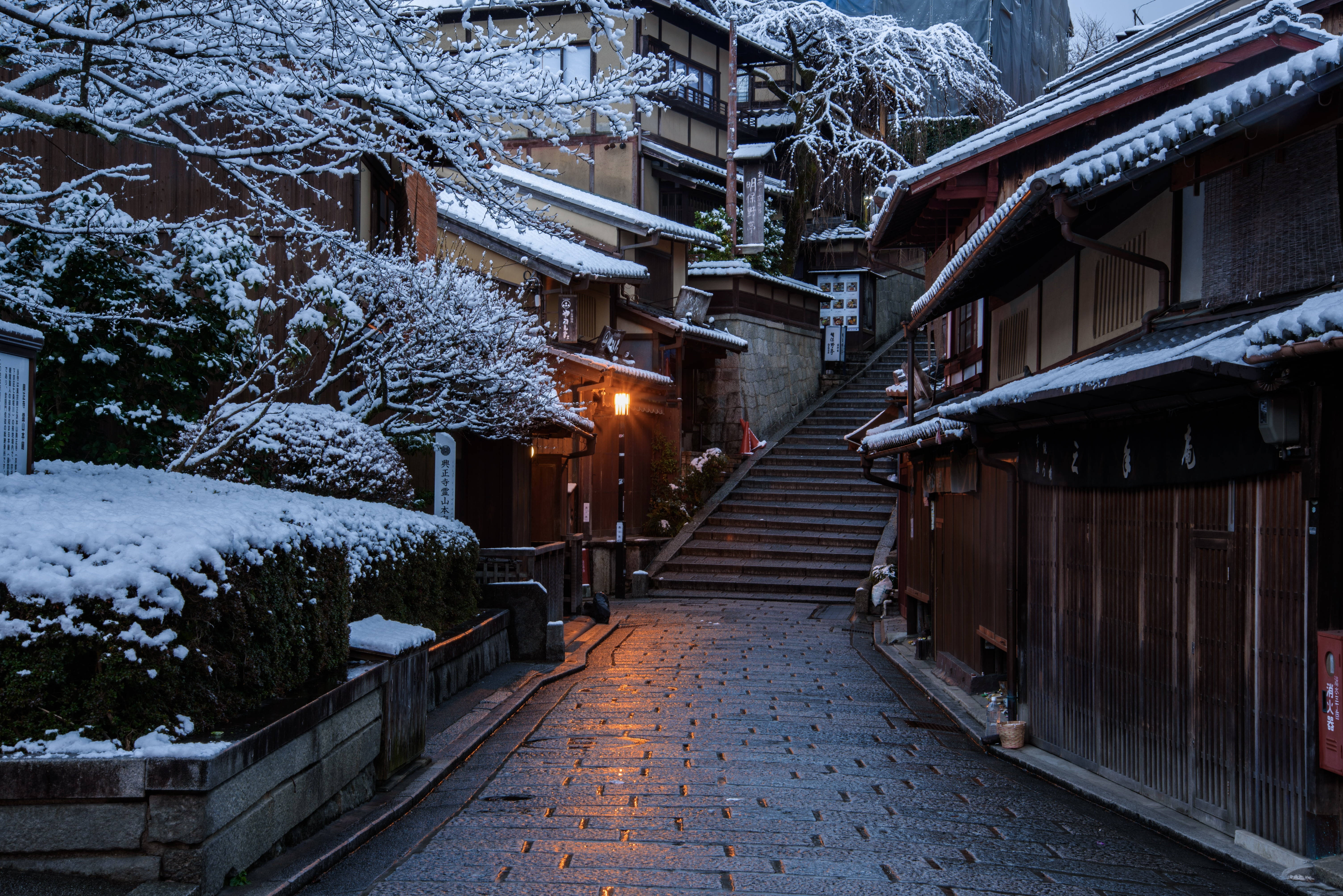 日本 桌布 壁紙 — 一條灑滿雪的傳統日本街道，夜晚溫暖的燈籠光線在濕滑的石階上反射。