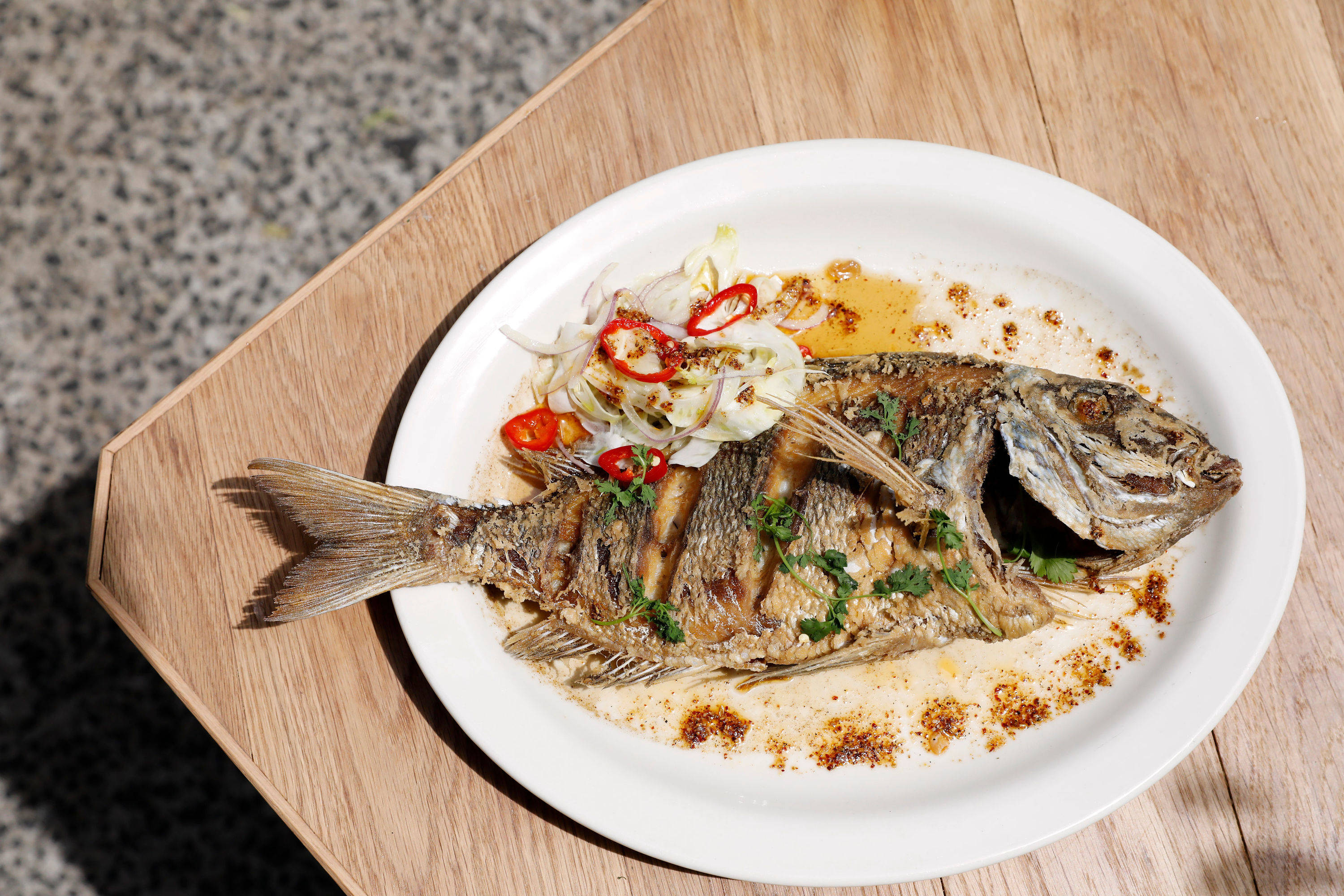 Seafood 桌布 壁紙 — 一條烤全魚配上紅辣椒和香草，放在白色盤子上，置於木托盤上，在明亮的陽光下。