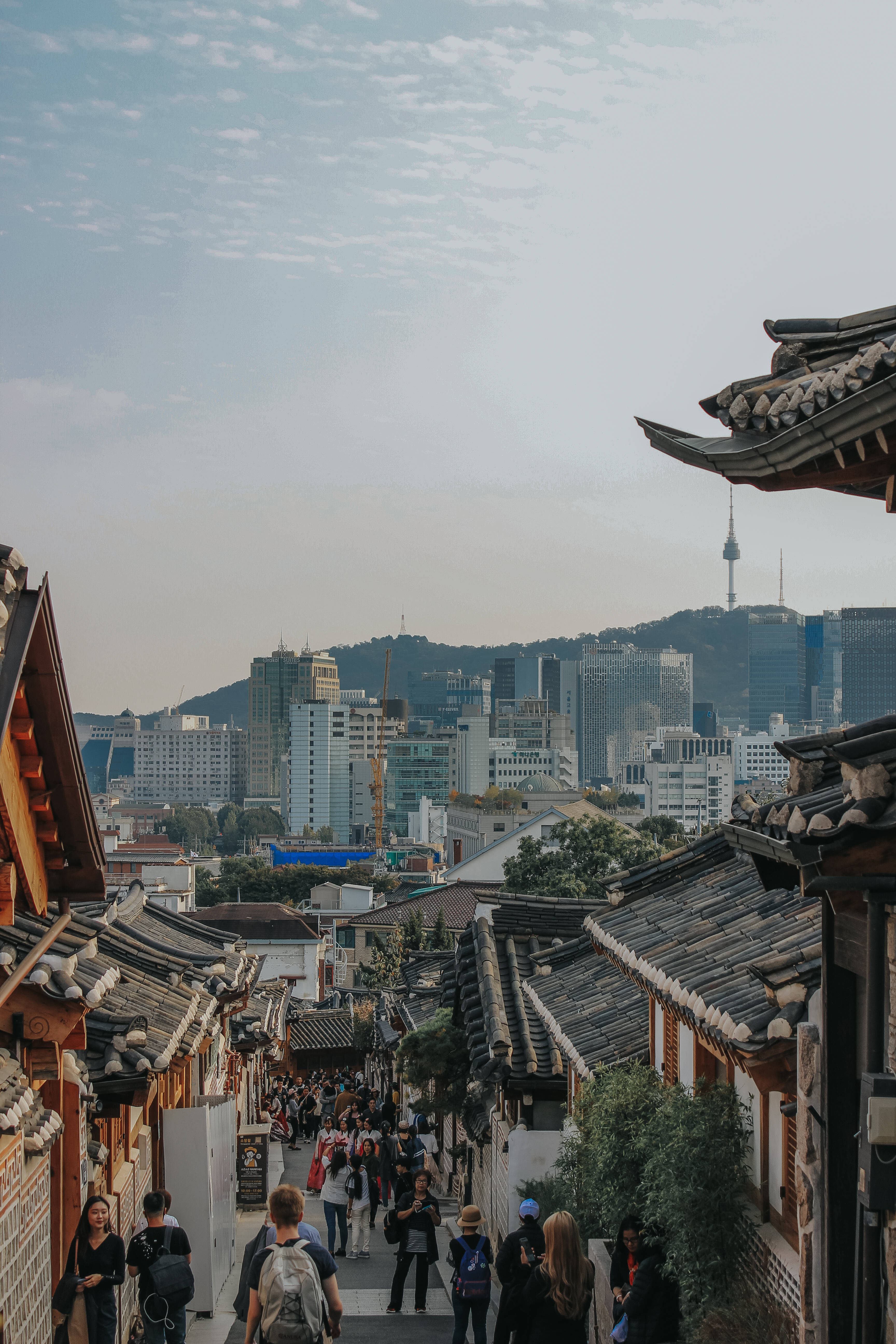 Seoul 桌布 壁紙 — 一條熱鬧狹窄的小巷，兩旁是傳統韓式瓦頂，通往現代城市天際線，背景是標誌性的N首爾塔，天空柔和多雲。