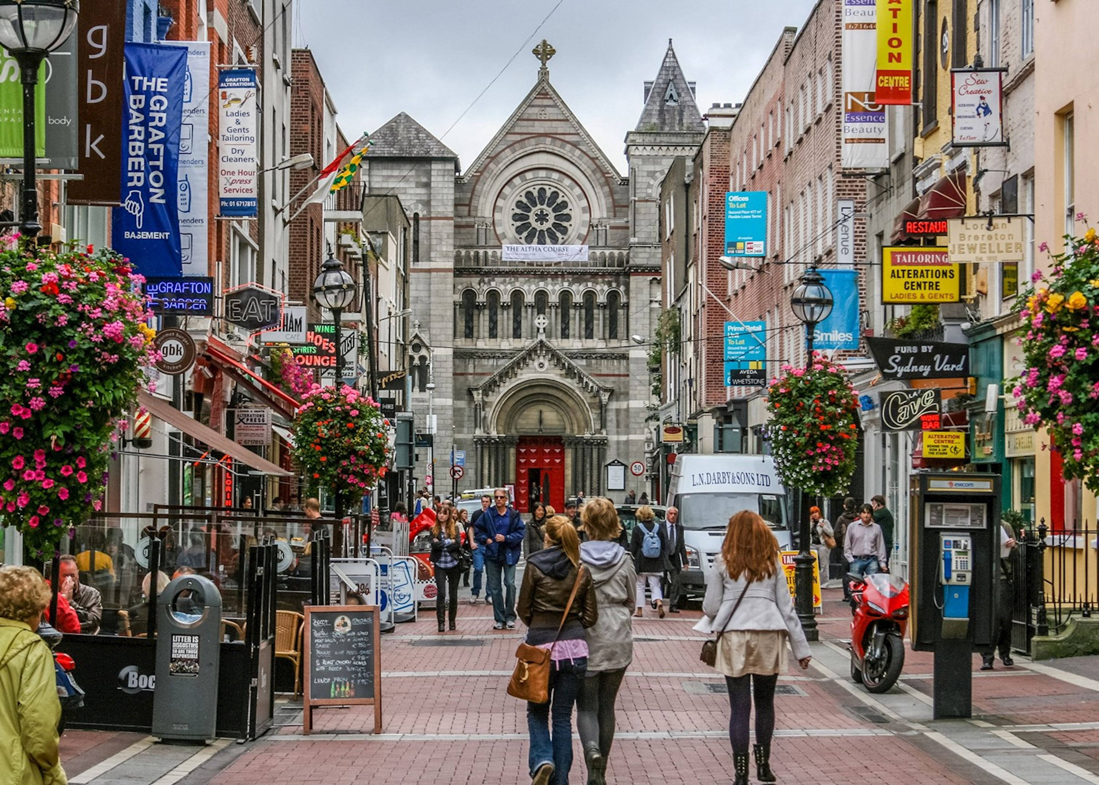 Europe 桌布 壁紙 — 一條熱鬧的行人街，兩旁是色彩繽紛的商店和懸掛的花籃，通往一座有紅色大門的宏偉石頭教堂。