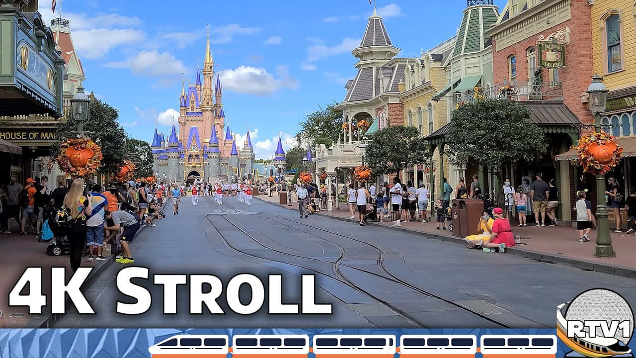 Disney World Desktop 桌布 壁紙 — 一條熱鬧的迪士尼主題公園街道，兩旁是色彩繽紛的建築物，人群在明亮的藍天下朝著宏偉的城堡前進。