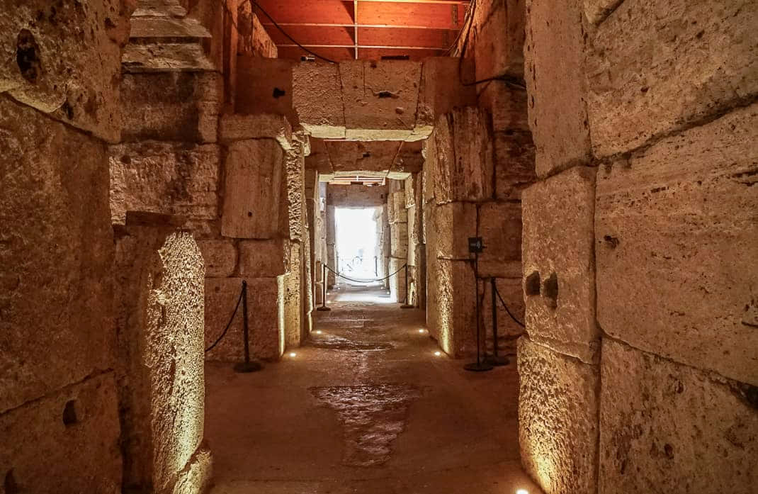 Roman Colosseum 桌布 壁紙 — 一條燈光昏暗的古老石頭走廊，溫暖的聚光燈照亮風化的石塊，通往明亮的出口。