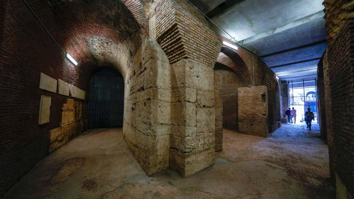 Roman Colosseum 桌布 壁紙 — 一條燈光昏暗的古老磚砌走廊，有拱形門口和石柱，由頭頂燈光照明，遠處可見人群。