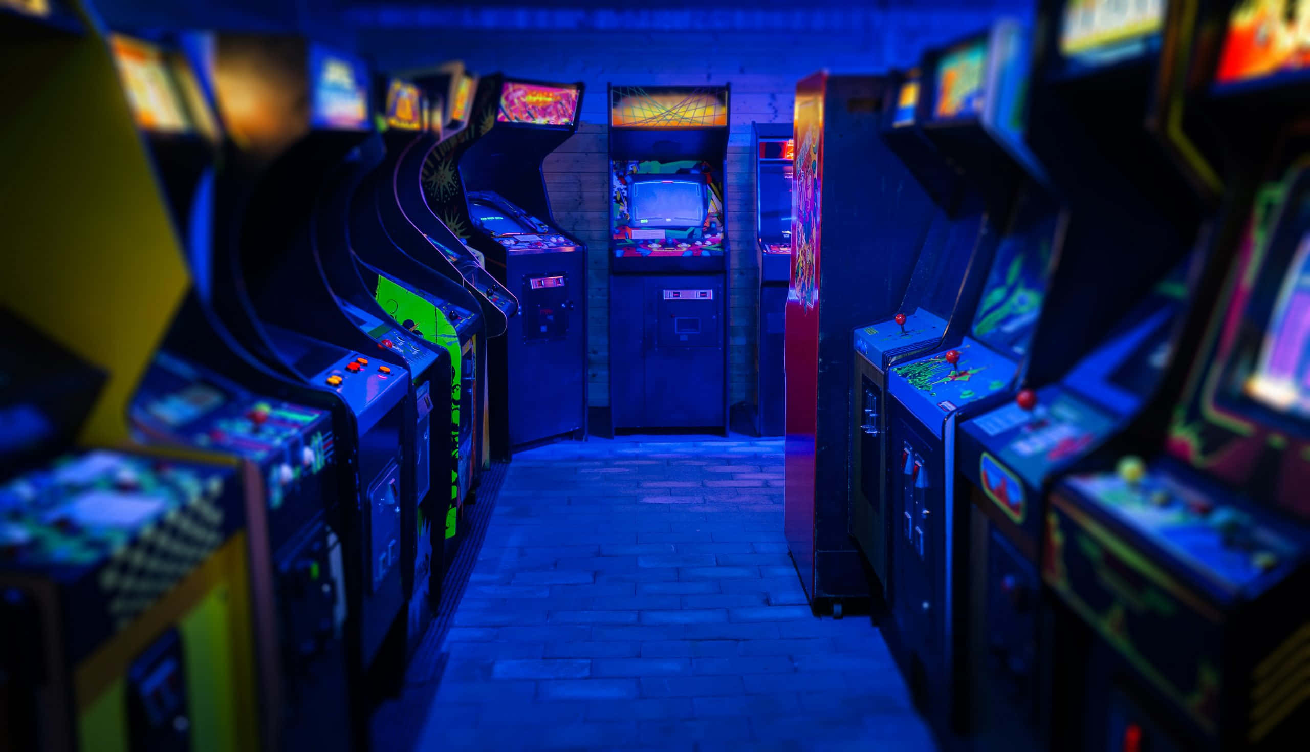 game 桌布 壁紙 — 一條燈光昏暗的拱廊通道，沐浴在藍色光芒中，兩旁排列著復古遊戲機。