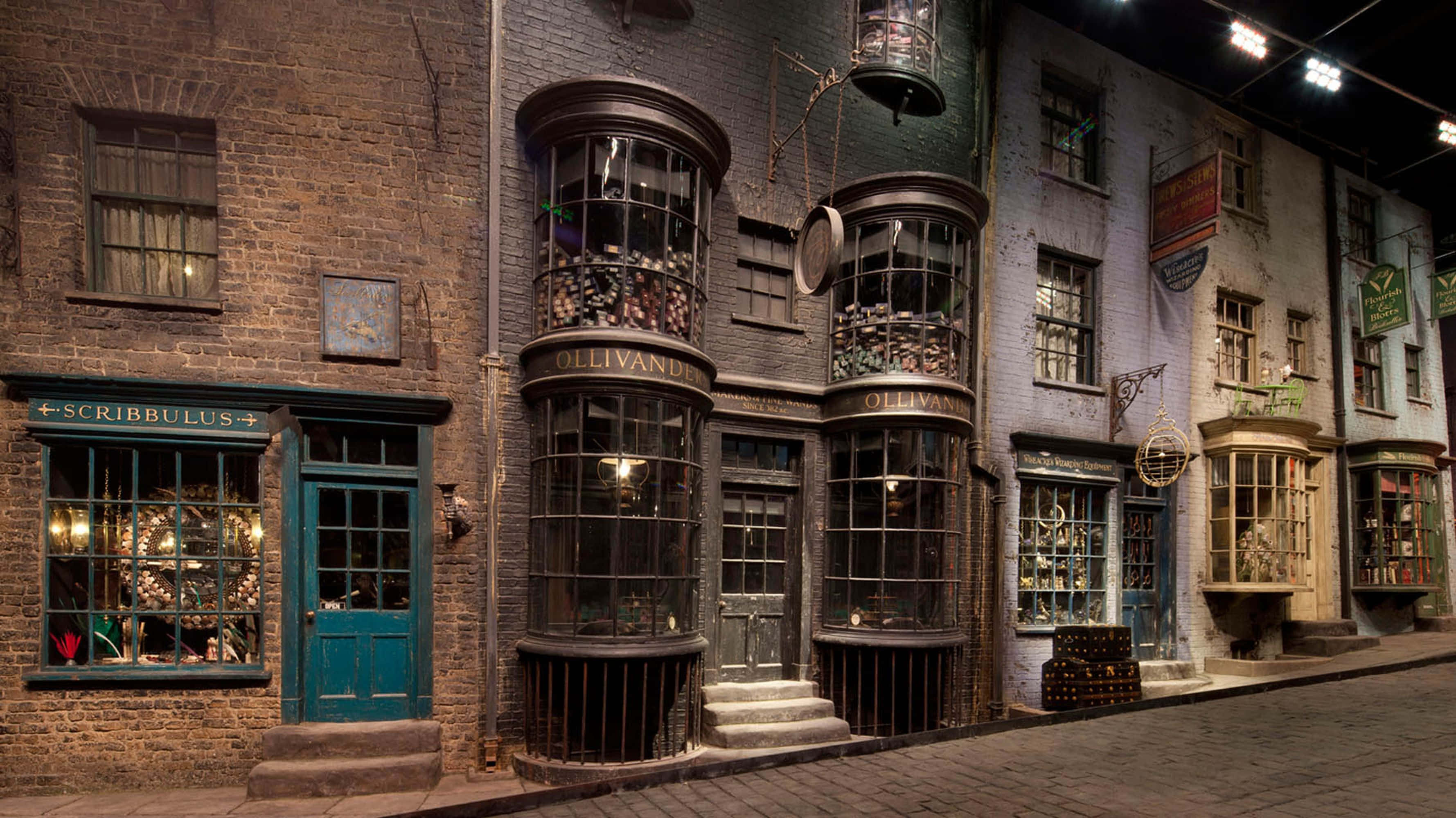 Diagon Alley 桌布 壁紙 — 一條燈光昏暗的鵝卵石街道，兩旁是奧利凡德和斯克里布勒斯等巫師商店，溫暖的燈光閃爍。
