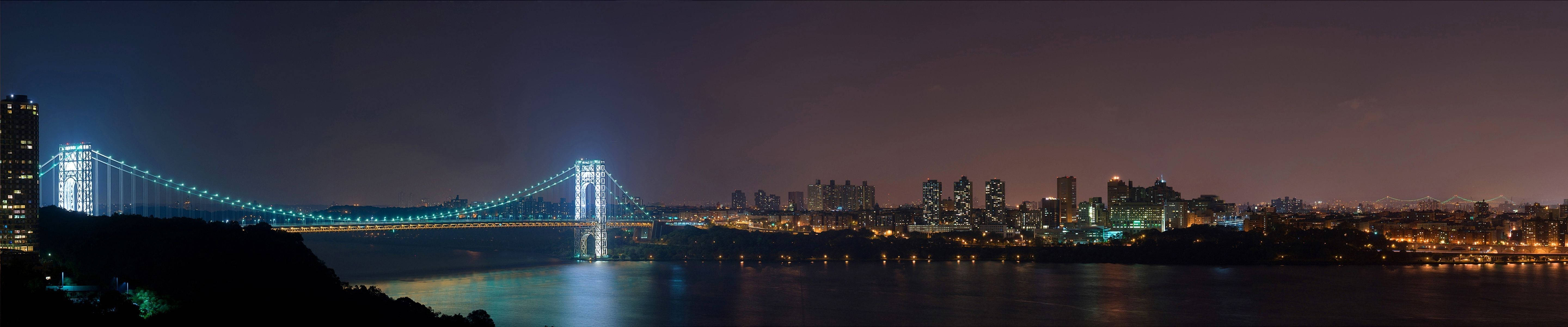 Triple 桌布 壁紙 — 一條燈火輝煌的吊橋橫跨黑暗河流的全景夜景，背景是閃爍的城市天際線。