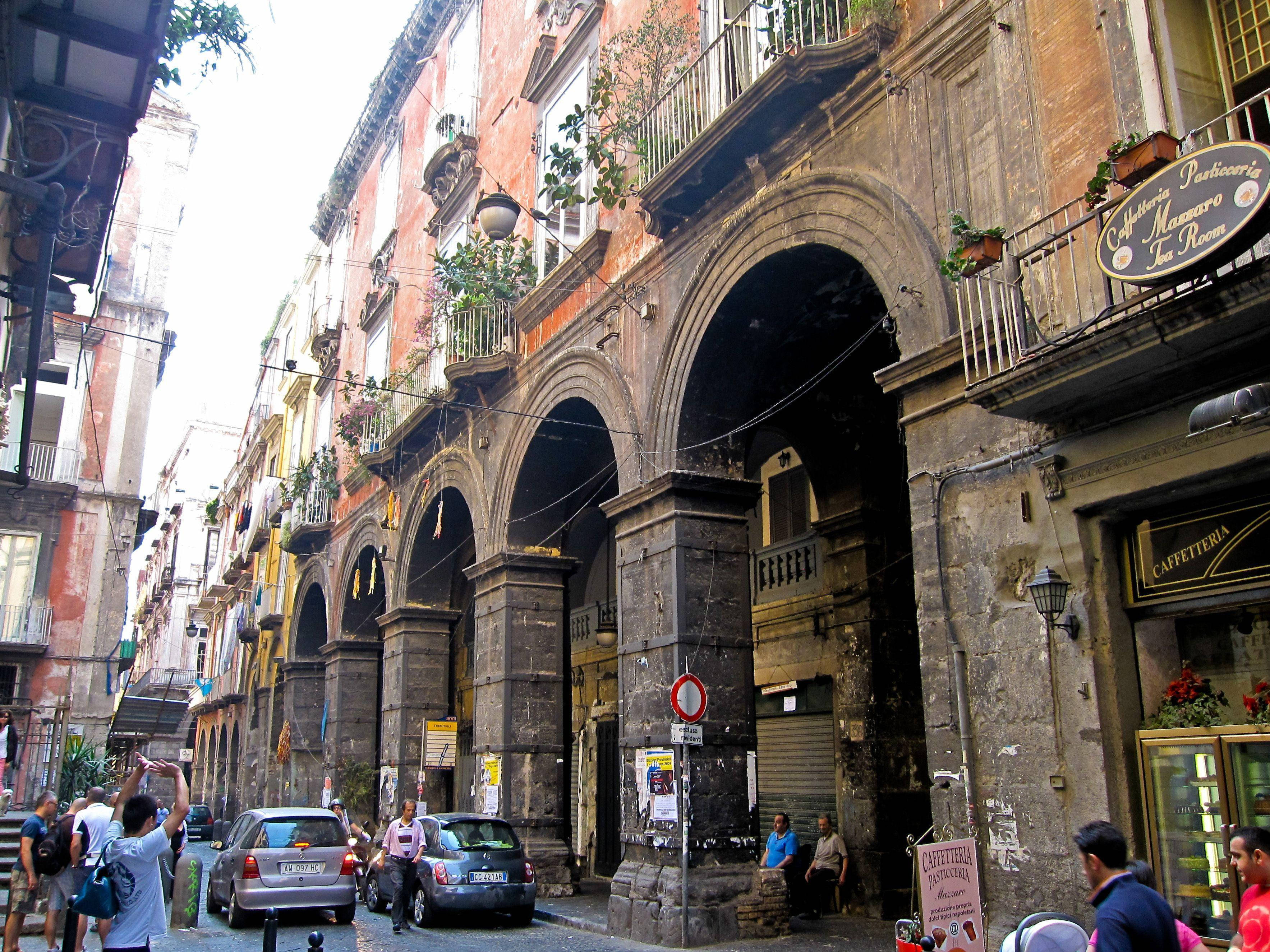 Naples 桌布 壁紙 — 一條狹窄、陽光明媚的街道，位於歷史悠久的歐洲城市，擁有拱形石柱廊、風化的建築物和行人。