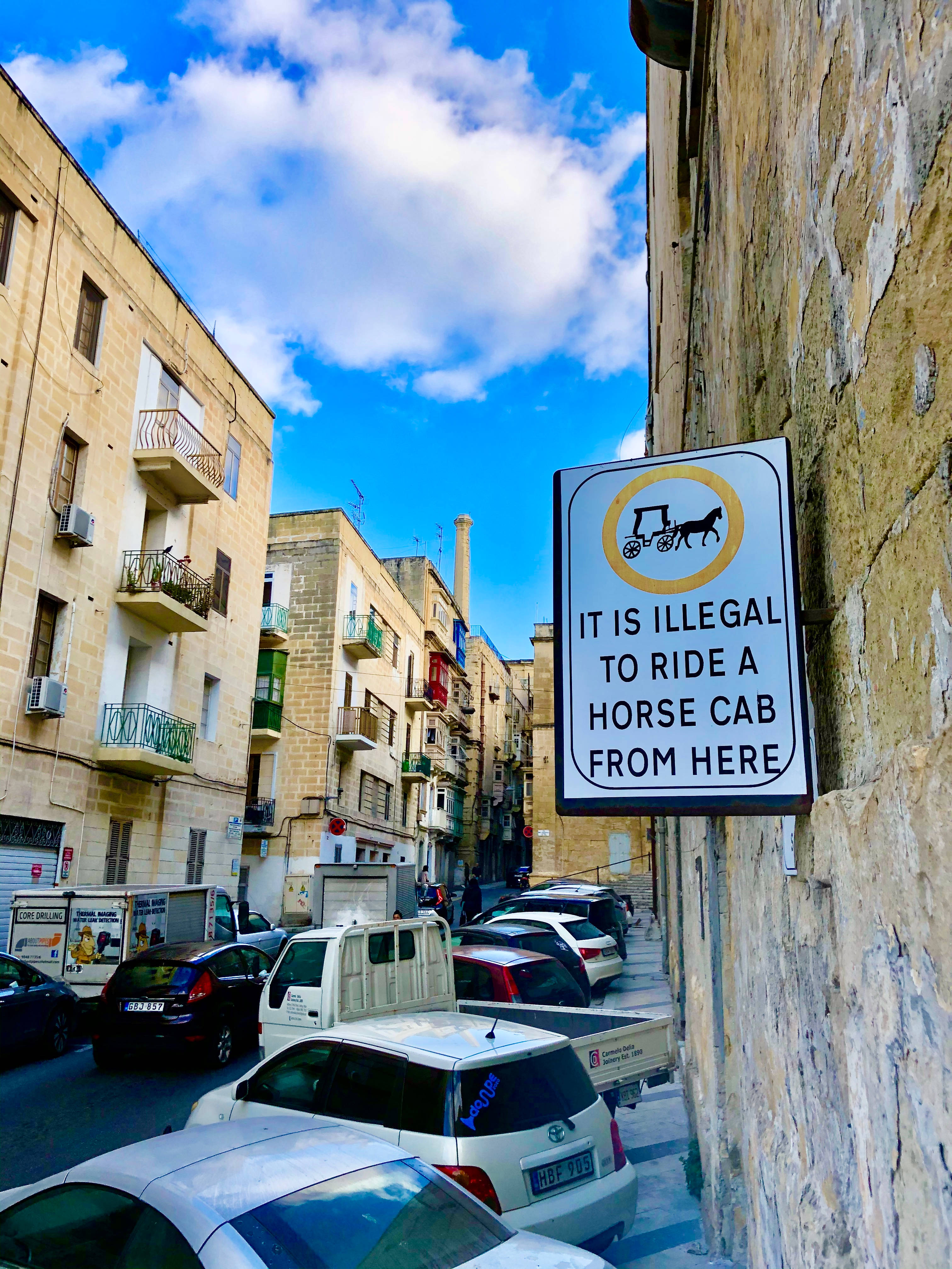 Malta 桌布 壁紙 — 一條狹窄、陽光明媚的街道，位於歷史悠久的歐洲小鎮，有停泊的汽車和一個禁止馬車的標誌，在明亮的藍天下。