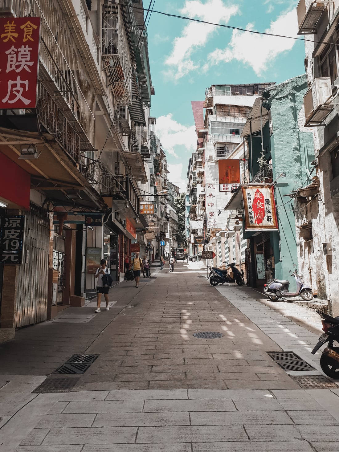 Chinatown 桌布 壁紙 — 一條狹窄、陽光普照的小巷，位於繁華的亞洲城市，兩旁有色彩繽紛的商店招牌和停放的機車。