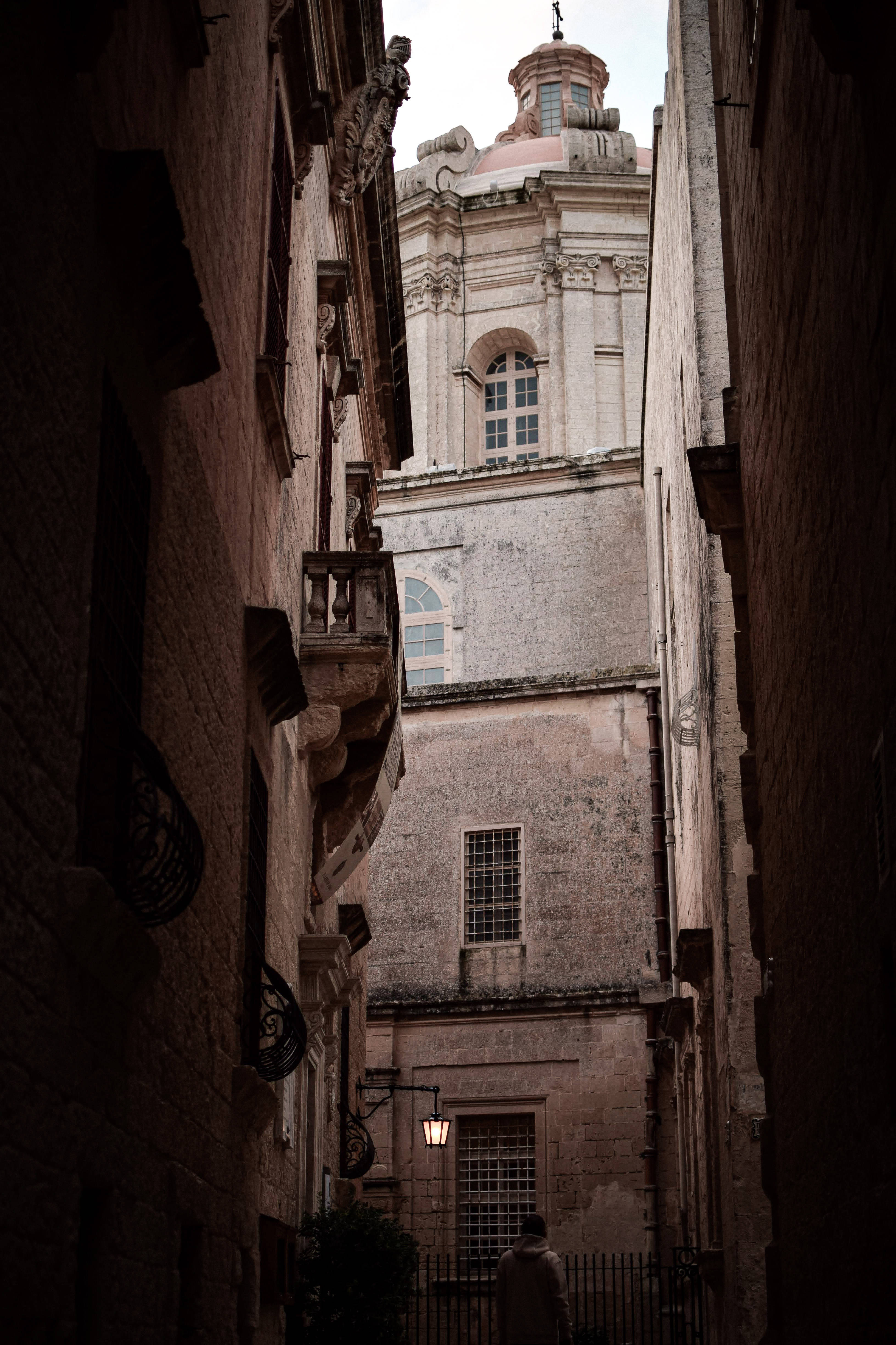 Malta 桌布 壁紙 — 一條狹窄、陽光普照的小巷，兩旁是古老的石造建築，盡頭顯露出一座宏偉的教堂塔樓高聳於屋頂之上。
