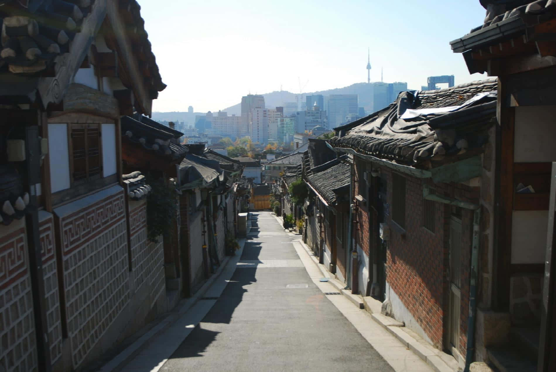 南韓 桌布 壁紙 — 一條狹窄、陽光普照的小巷，兩旁是傳統韓式瓦頂房屋，通向遠處有高塔的現代城市天際線。