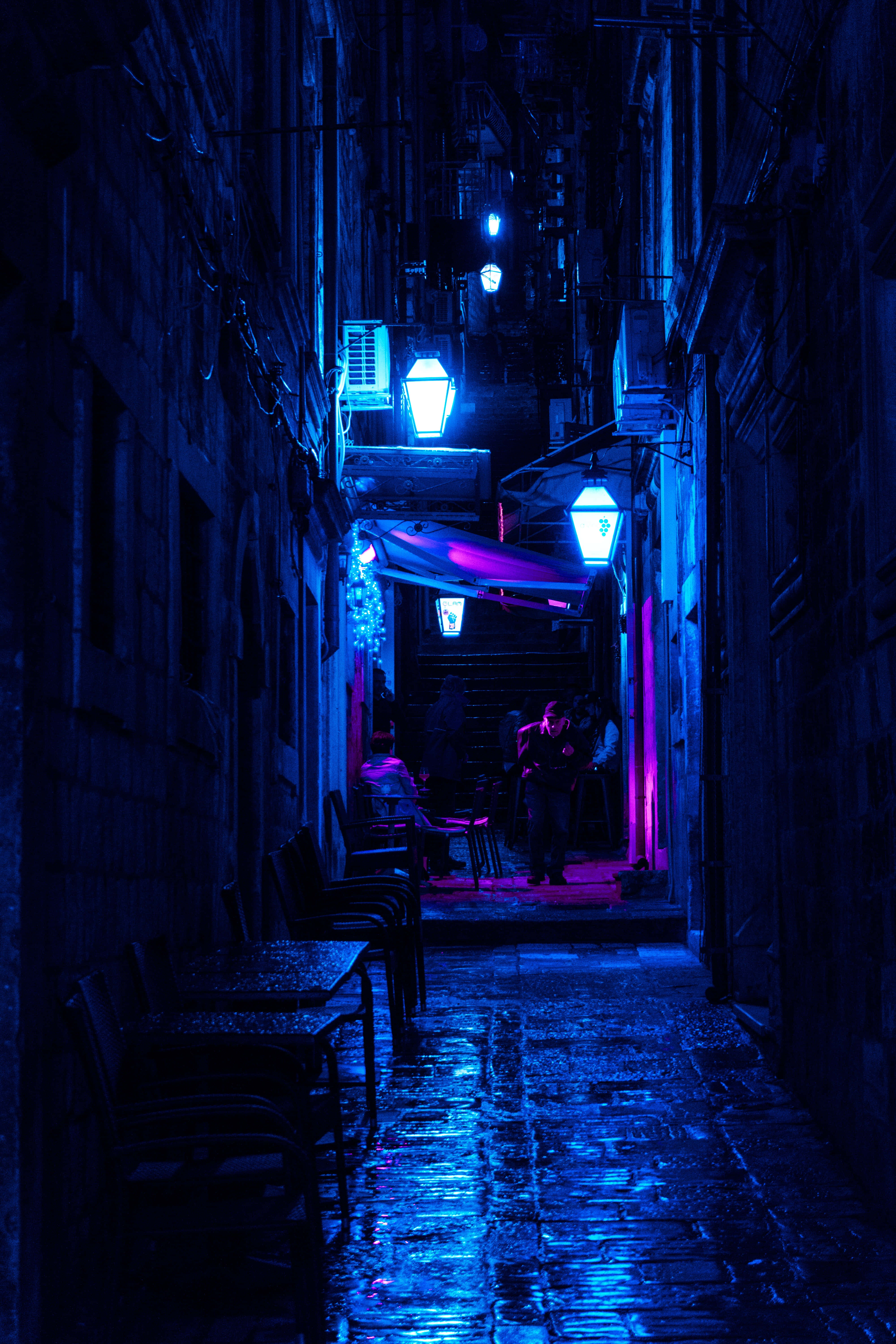 Night Street 桌布 壁紙 — 一條狹窄潮濕的小巷在夜晚沐浴在電光藍色霓虹燈中，燈光反射在鵝卵石上，遠處的咖啡館有剪影人物。