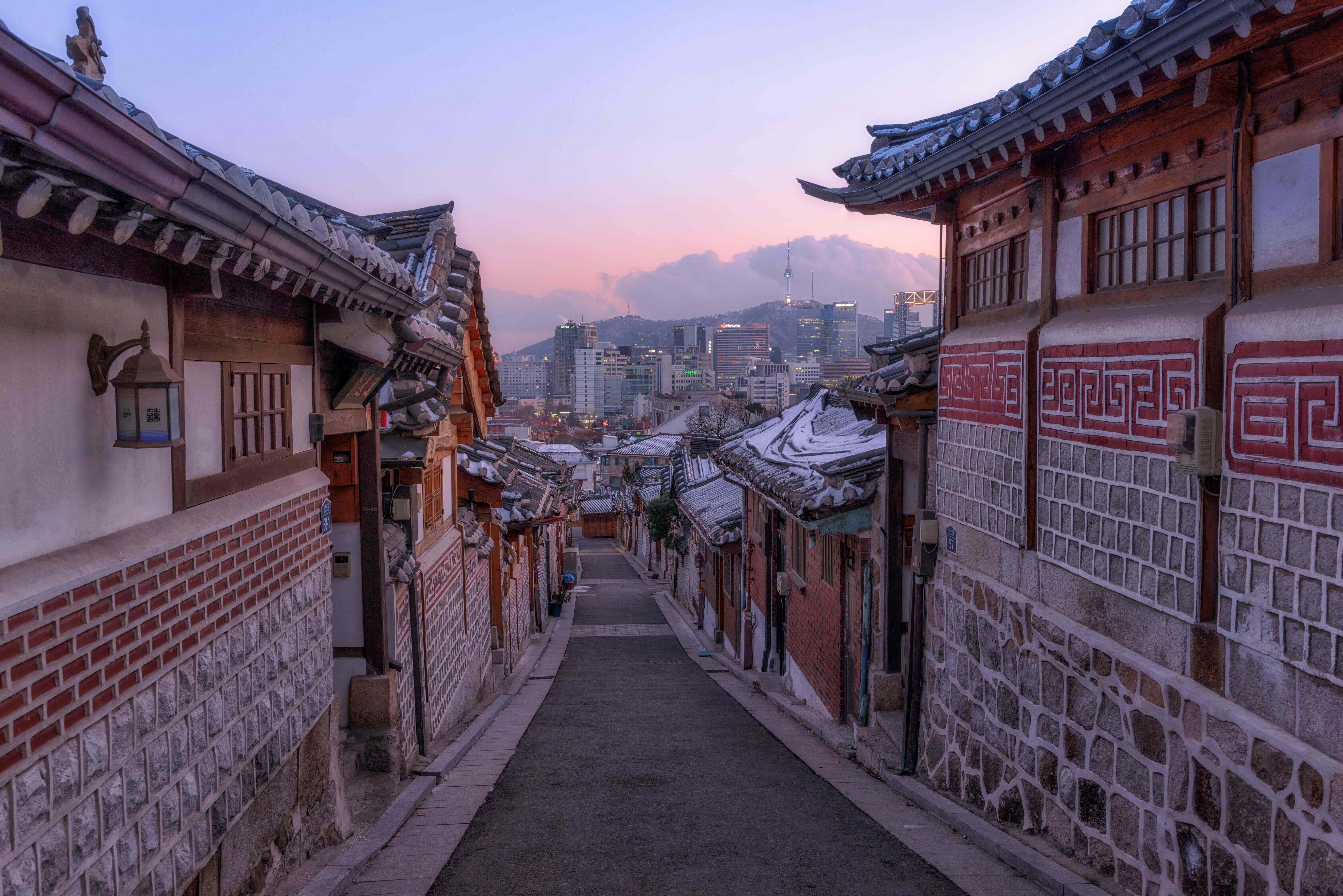 韓國 桌布 壁紙 — 一條狹窄的傳統韓國小巷，兩旁是石頭和瓷磚建築，向上延伸至黃昏時分的現代城市天際線。