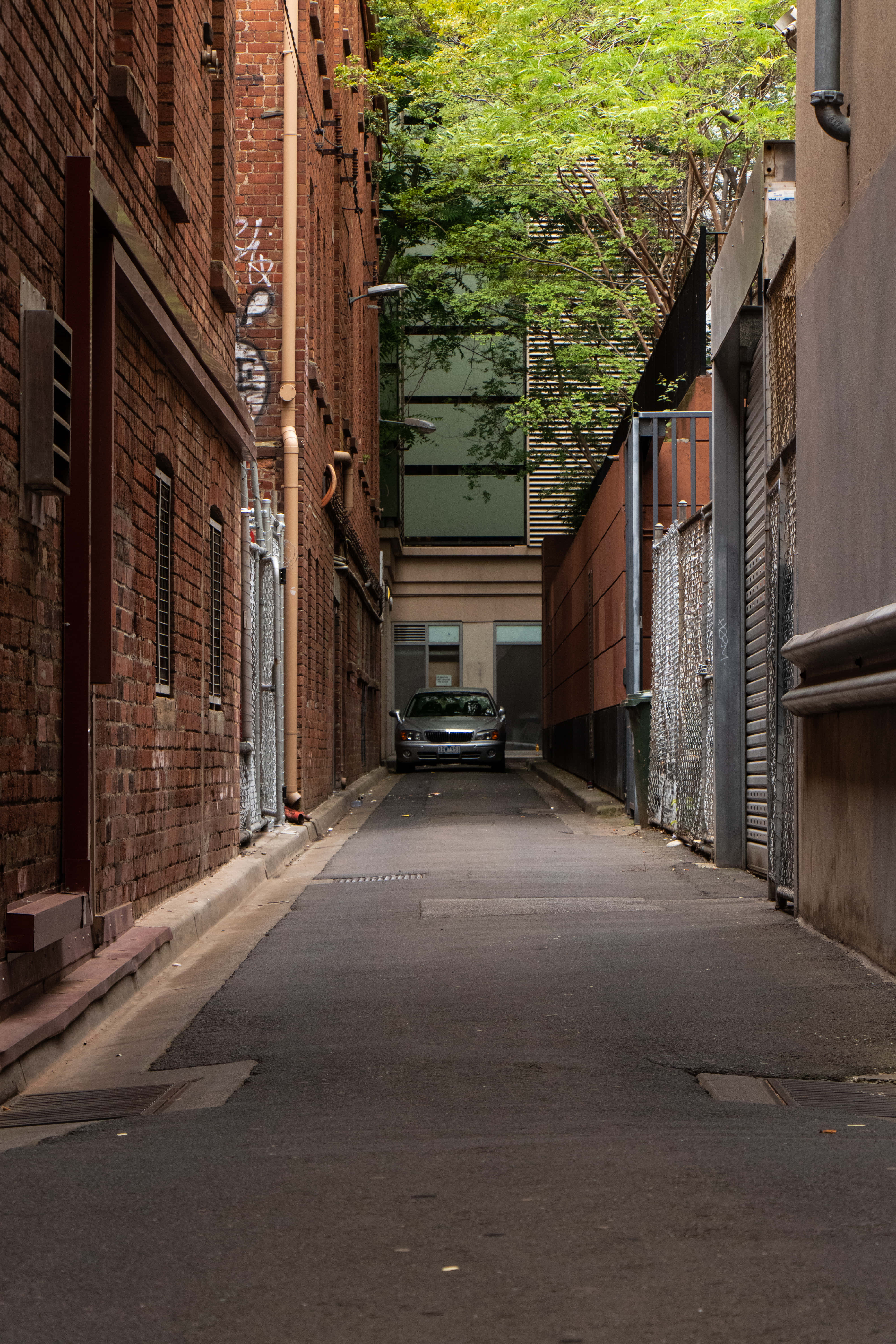 Alleyway 桌布 壁紙 — 一條狹窄的城市小巷，兩旁是磚砌建築和金屬百葉窗，盡頭有一輛銀色汽車停在綠色樹葉下。