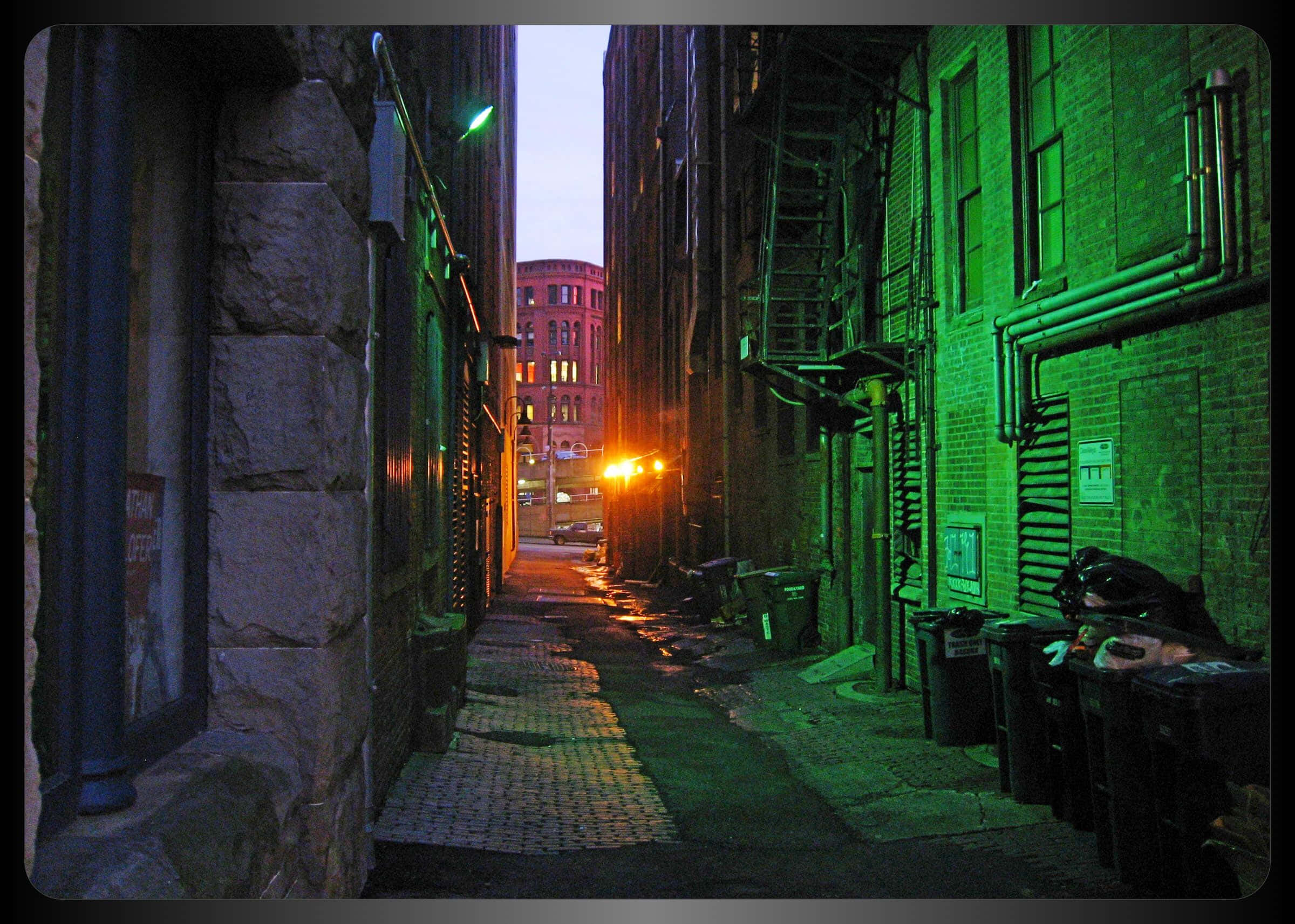 Alleyway 桌布 壁紙 — 一條狹窄的城市小巷，沐浴在詭異的綠光中，遠處磚造建築在黃昏時散發出溫暖的光芒。