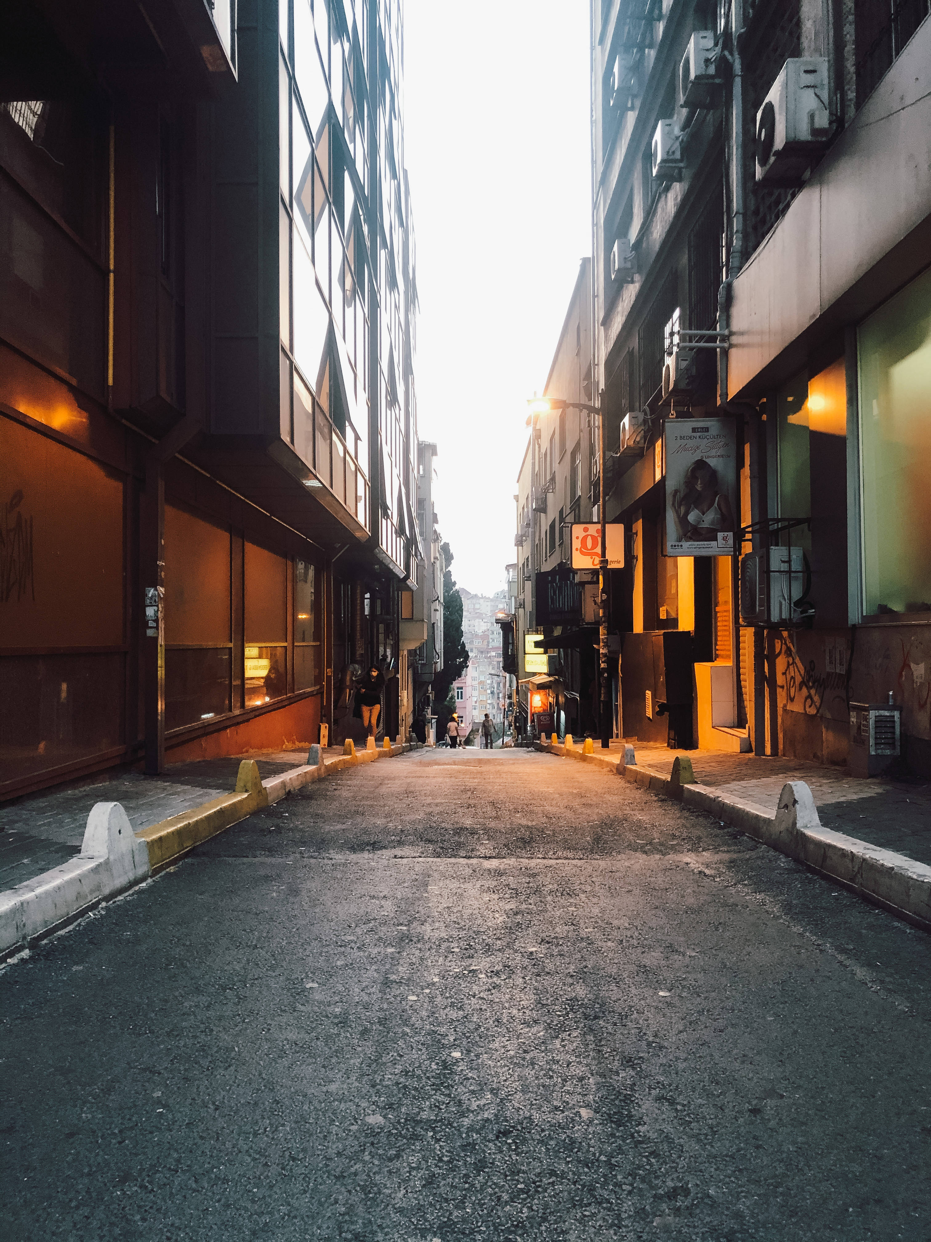 Street 桌布 壁紙 — 一條狹窄的城市小巷在高樓之間向上延伸，溫暖的燈光閃爍，遠處有行人，天空明亮。