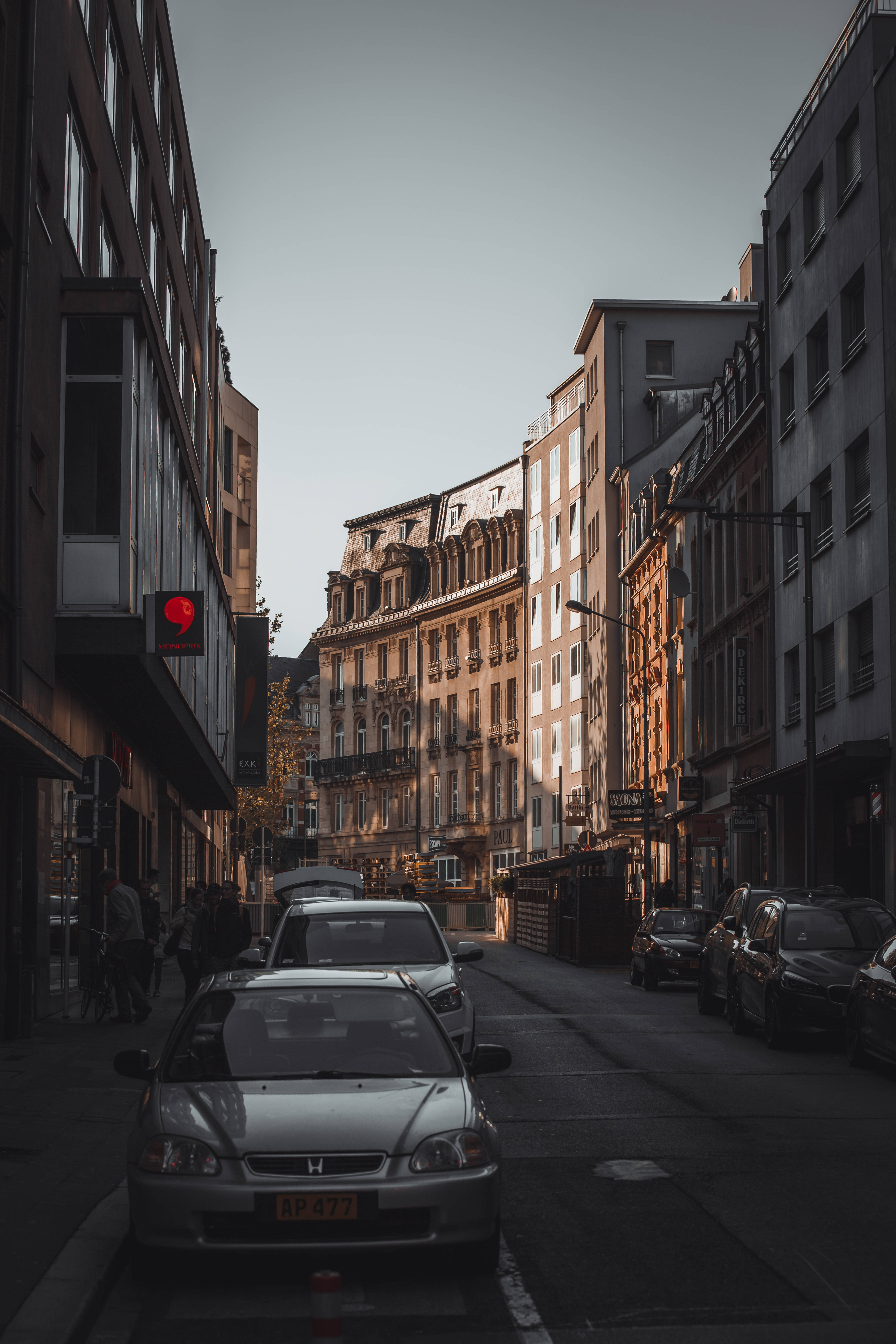Luxembourg 桌布 壁紙 — 一條狹窄的城市街道，兩旁停滿汽車和高樓大廈，沐浴在柔和的金色時刻光線中。