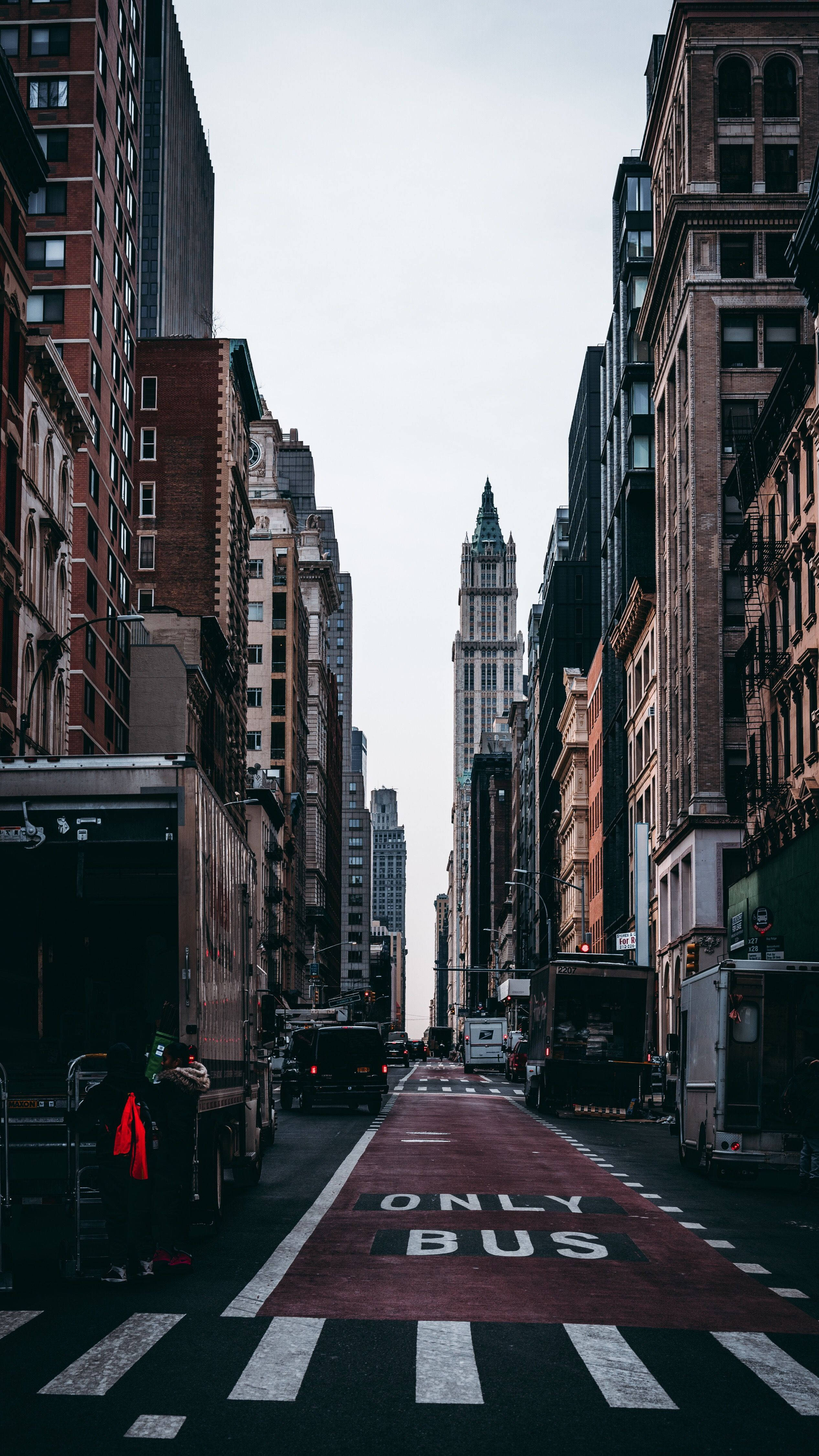 new york city iPhone x 桌布 壁紙 — 一條狹窄的城市街道，兩旁是高聳的磚造建築，一條紅色公車專用道在陰天中通往遠處的摩天大樓。