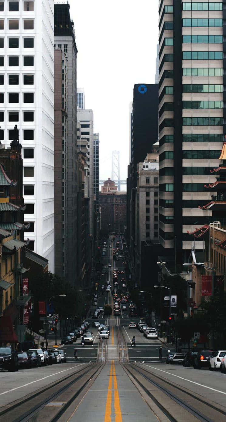 城市 桌布 壁紙 — 一條狹窄的城市街道，兩旁是高聳的摩天大樓和歷史建築，電車軌道在陰天之下通往遠處的一座橋樑。