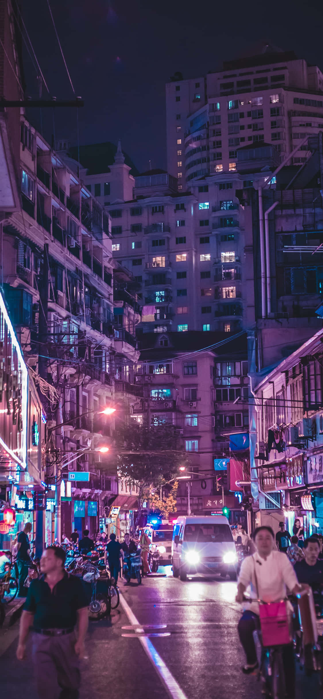 iPhone 桌布 壁紙 — 一條狹窄的城市街道在夜晚被建築物和商店櫥窗的霓虹紫色和藍色燈光所籠罩，人們和車輛穿梭其中。