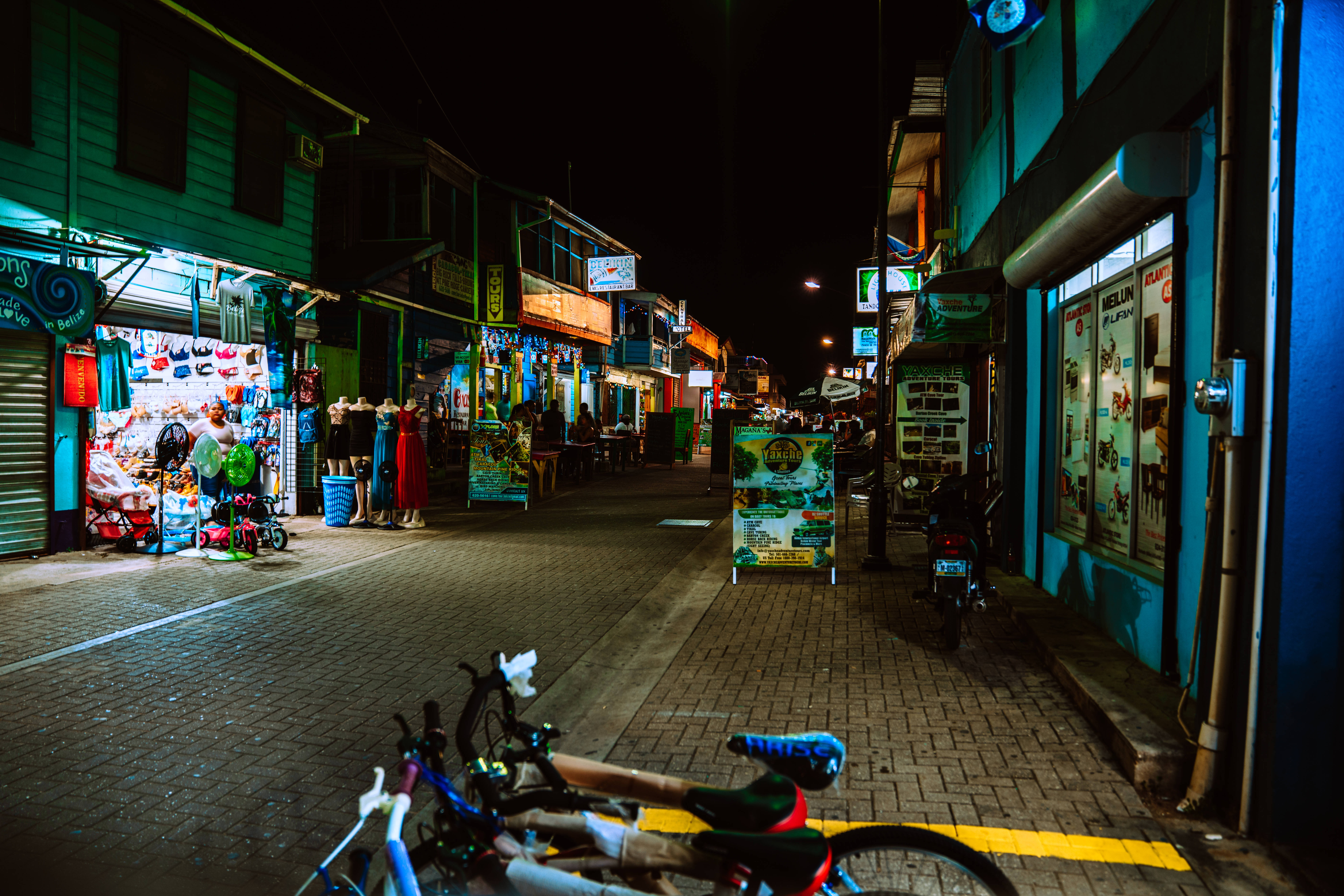 Belize 桌布 壁紙 — 一條狹窄的夜間街道，兩旁是燈火通明的商店和停放的自行車，天空漆黑一片。