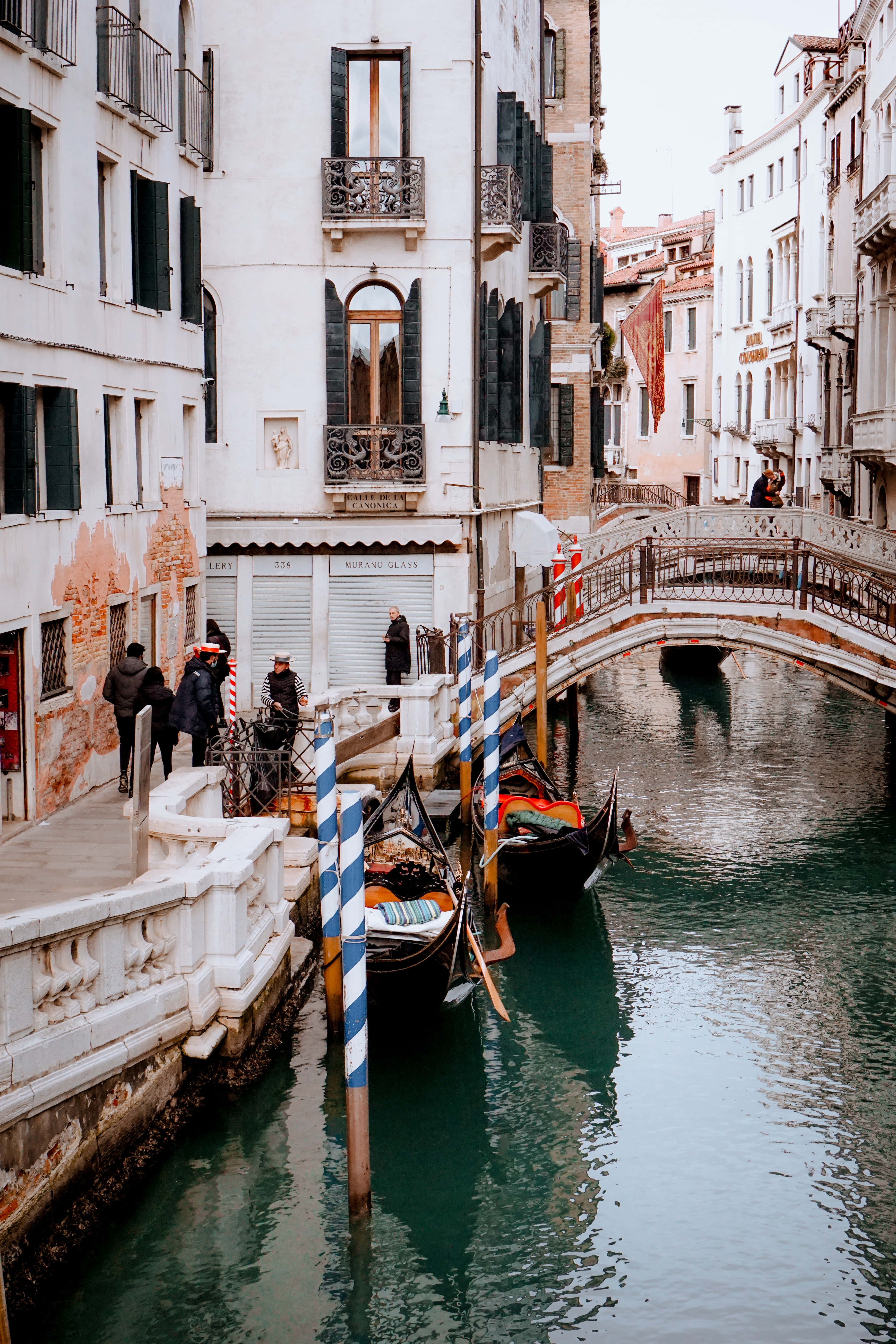Italy Aesthetic 桌布 壁紙 — 一條狹窄的威尼斯運河，有貢多拉停泊在石橋旁，以及帶綠色百葉窗的歷史建築。