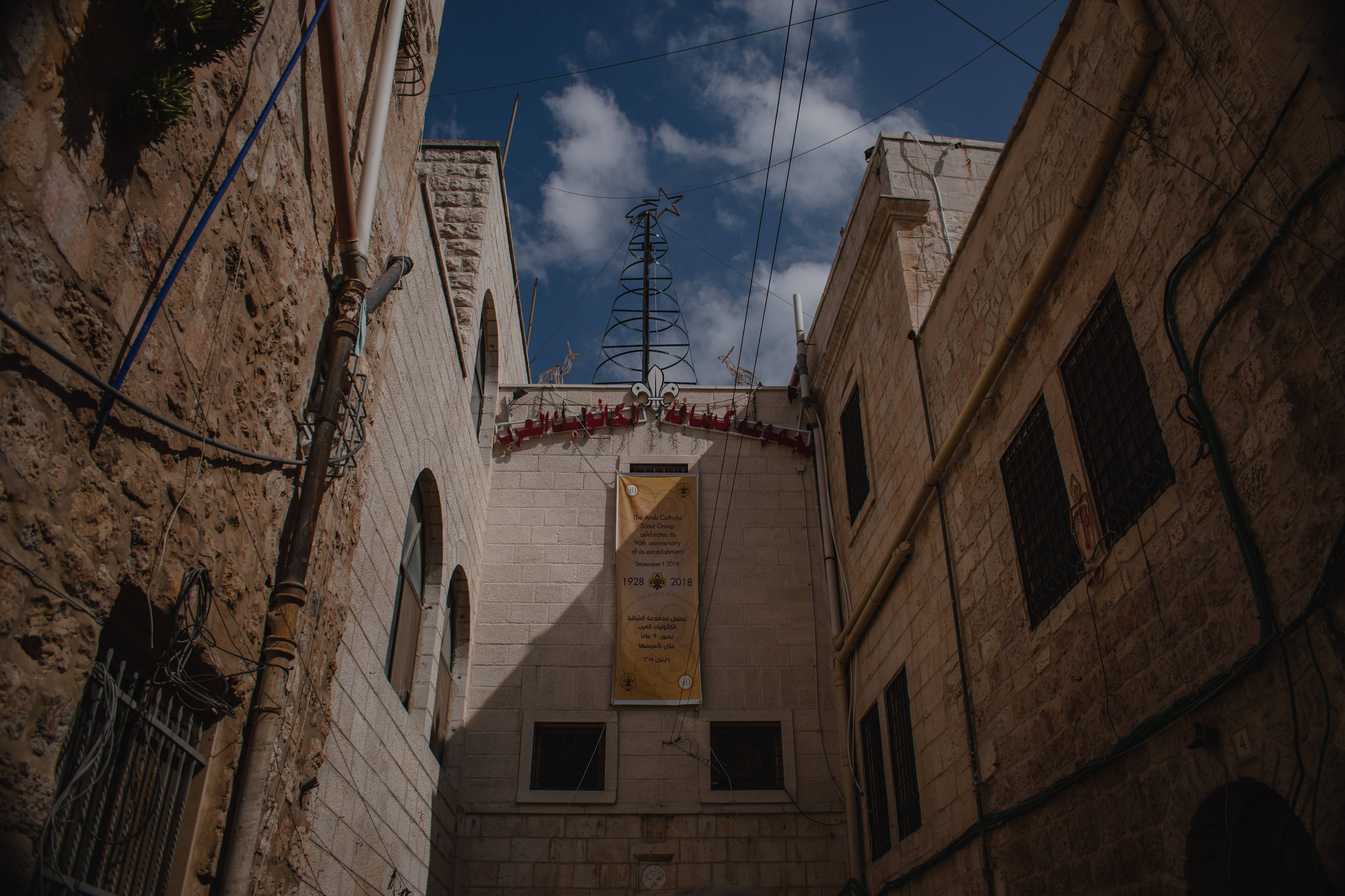 Israel 桌布 壁紙 — 一條狹窄的小巷，兩旁是風化的石頭建築，通向明亮的藍天，有一個金屬樹雕塑和懸掛的橫幅。