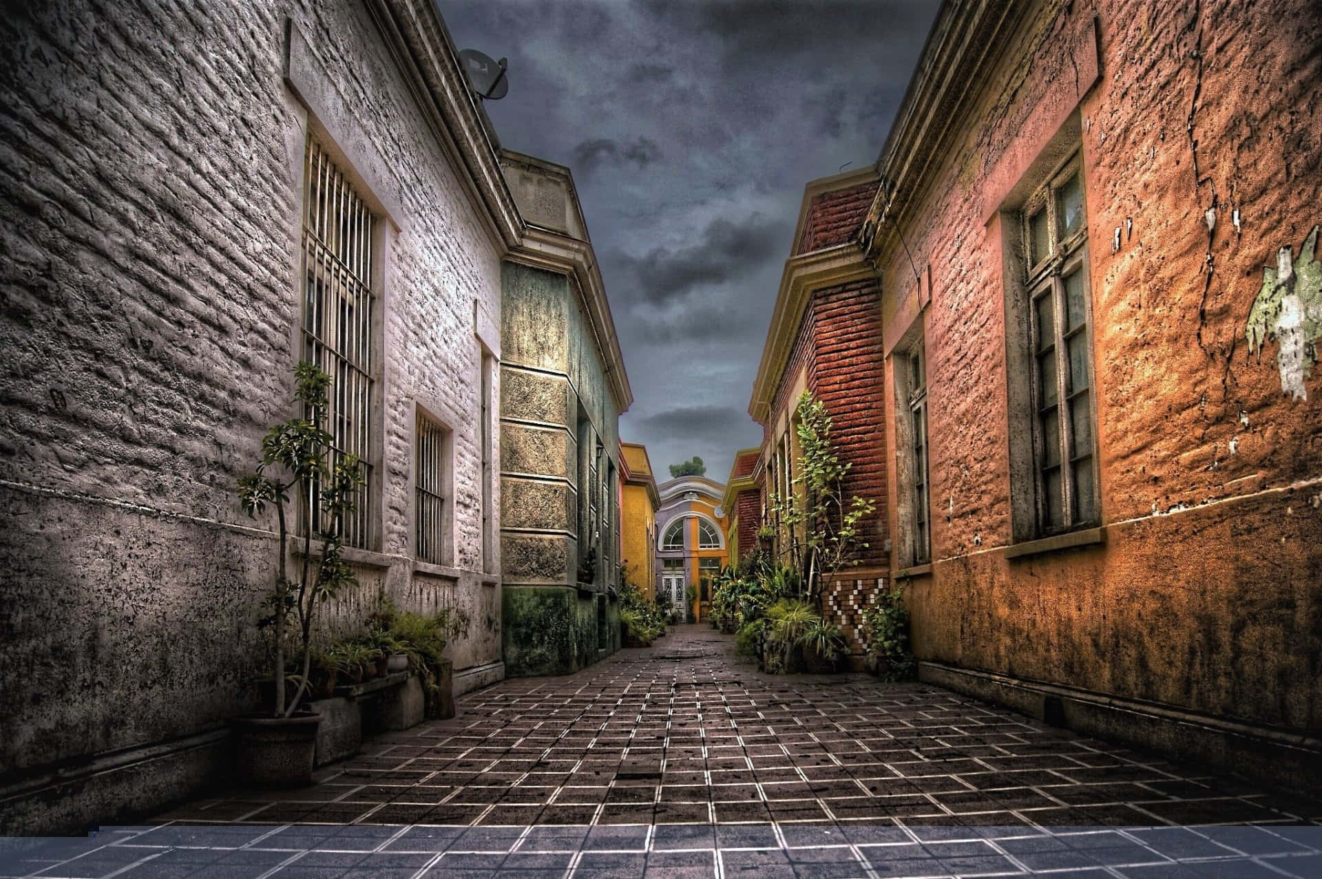 Alleyway 桌布 壁紙 — 一條狹窄的小巷，兩旁是風化的磚造建築，在暴風雨的天空下，盆栽植物排列在鵝卵石小徑上。