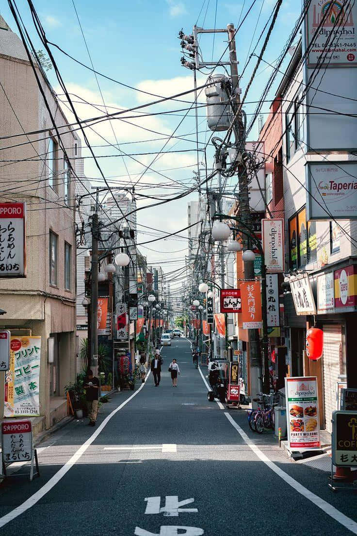 Japanese Phone 桌布 壁紙 — 一條狹窄的日本街道，兩旁掛滿色彩繽紛的商店招牌和纏繞的架空電線，上方是明亮的藍天。