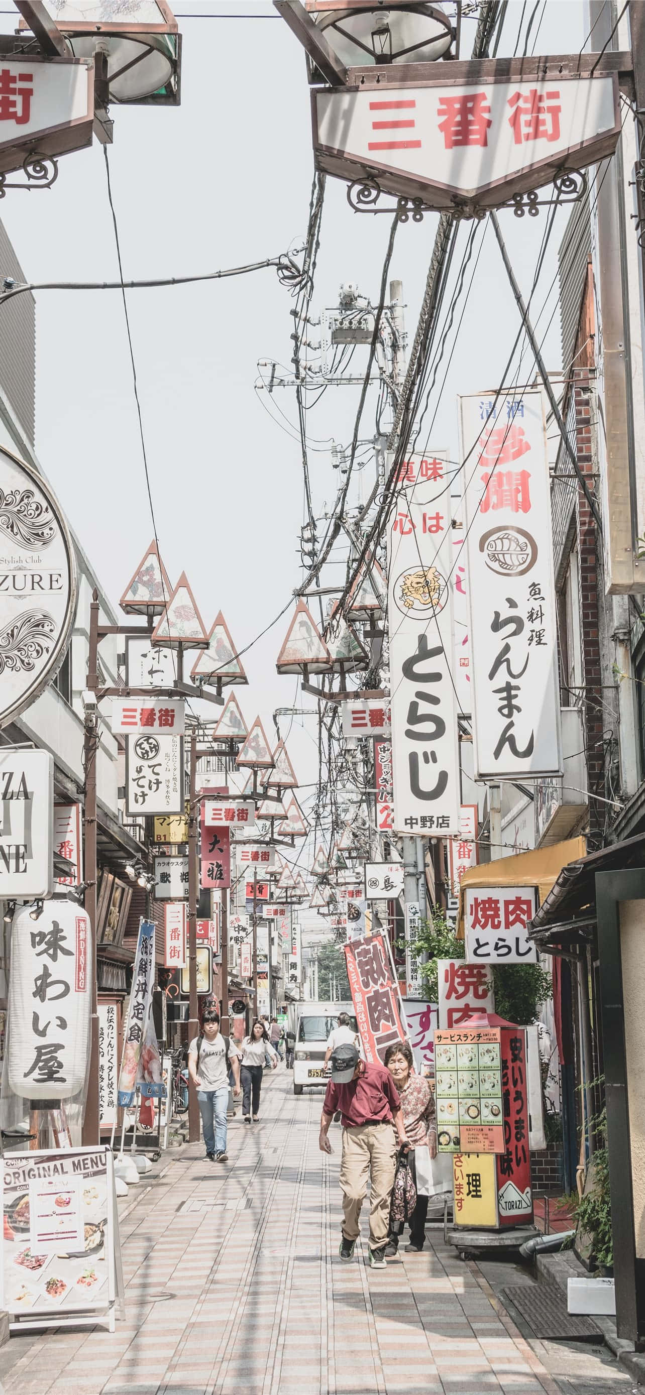 Japanese iPhone 桌布 壁紙 — 一條狹窄的日本街道，掛滿招牌，行人在明亮的天空下行走。