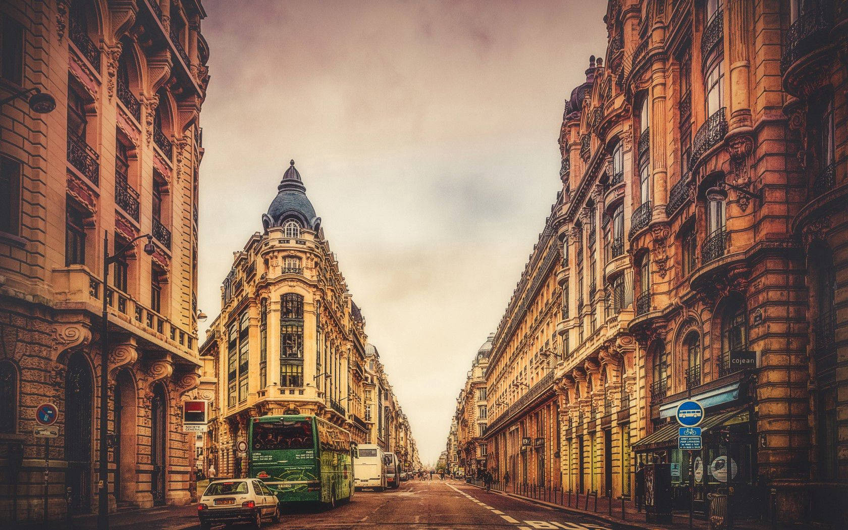 France 桌布 壁紙 — 一條狹窄的歐洲街道，兩旁是華麗的歷史建築，在柔和的多雲天空下，有一輛綠色巴士和汽車沿路邊停放。