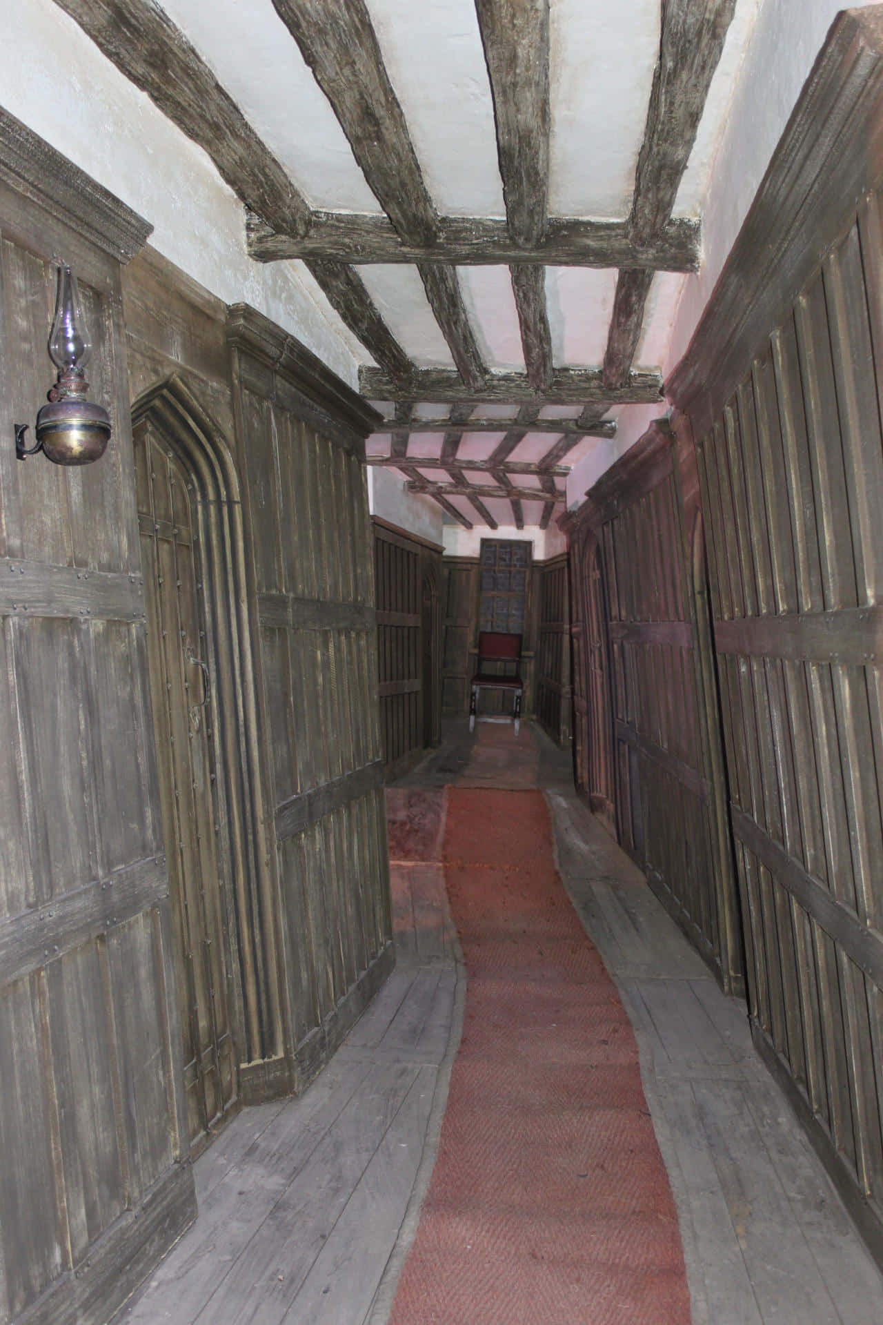 The Hogwarts Charms Corridor 桌布 壁紙 — 一條狹窄的歷史木製走廊，有外露橫樑和紅色地毯，通向遠處的椅子。