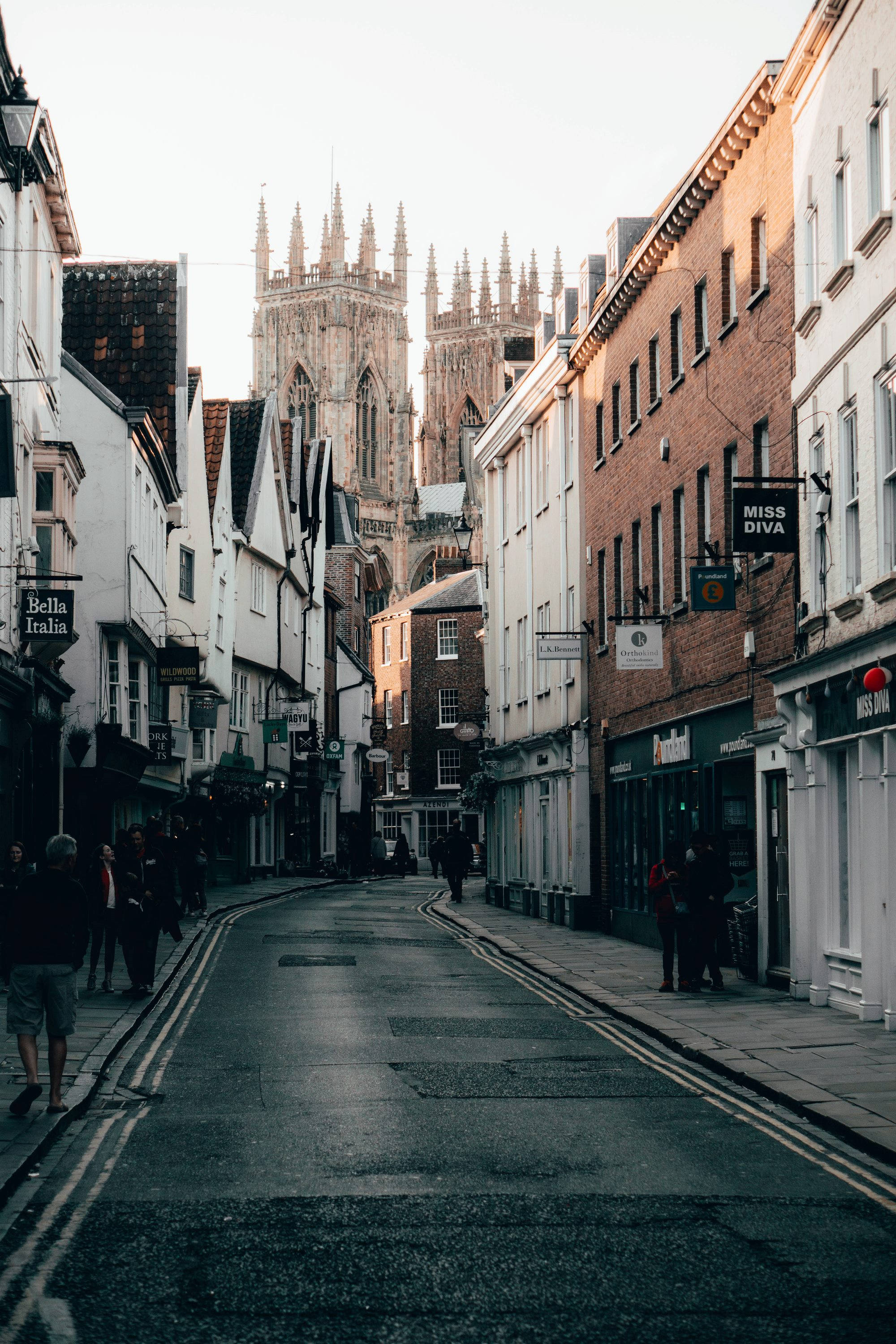 England 桌布 壁紙 — 一條狹窄的歷史街道，兩旁是磚砌和白色的建築，通往一座在柔和天空下高聳的哥德式大教堂。