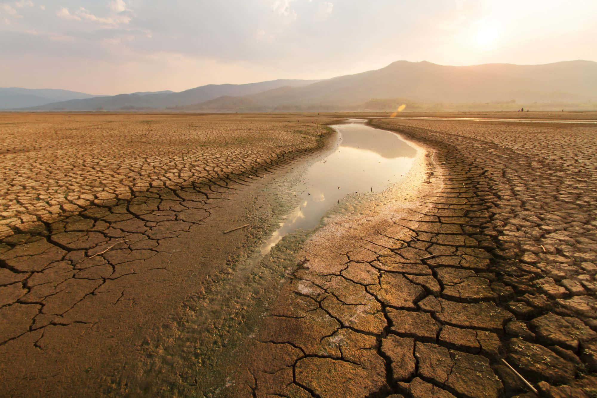 climate change 桌布 壁紙 — 一條狹窄的水流蜿蜒穿過廣闊的龜裂土地，背景是朦朧的日落天空和遠山。