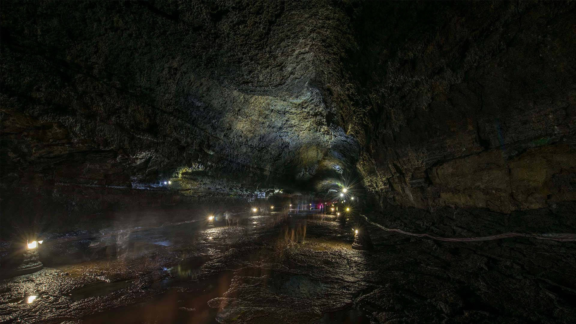 Jeju 桌布 壁紙 — 一條狹窄的洞穴，由人造燈光照亮，光線在濕岩石上反射，遊客沿著小路行走。