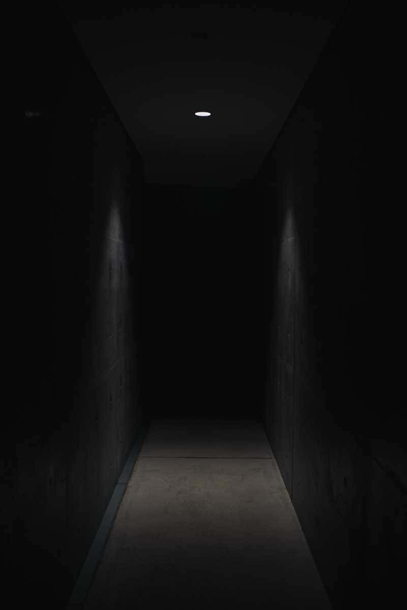 Dark Corner 桌布 壁紙 — 一條狹窄的混凝土走廊陷入黑暗，僅由一盞天花板聚光燈照亮，投下長長的陰影。