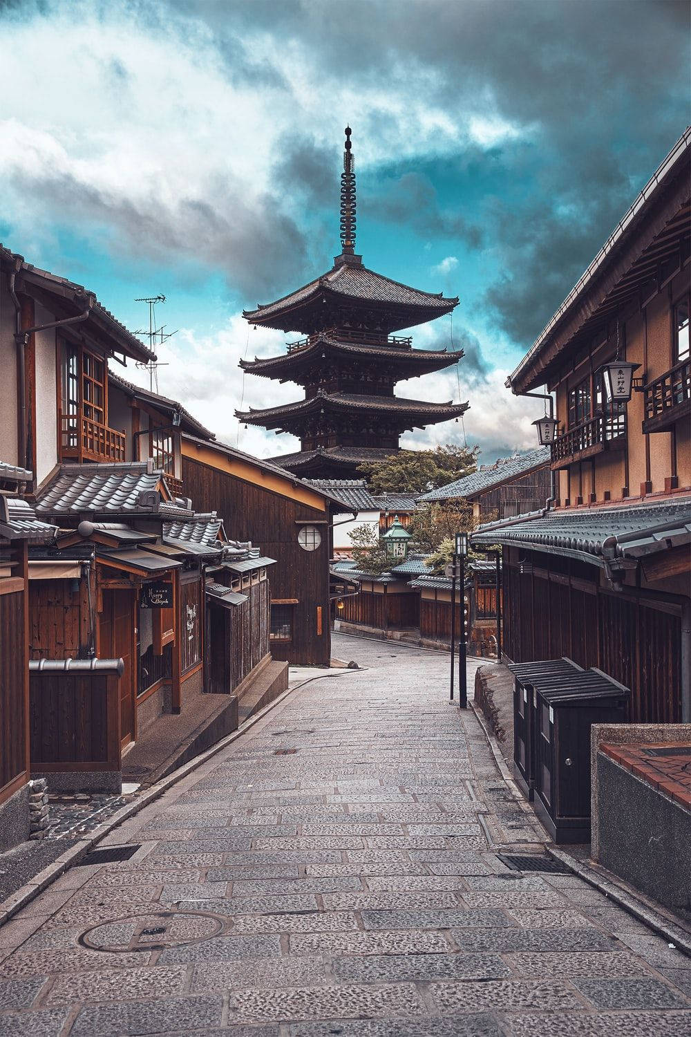 京都 桌布 壁紙 — 一條狹窄的石板街道在傳統日本小鎮中，通往一座高聳的木造寶塔，天空烏雲密布，氣氛戲劇化。