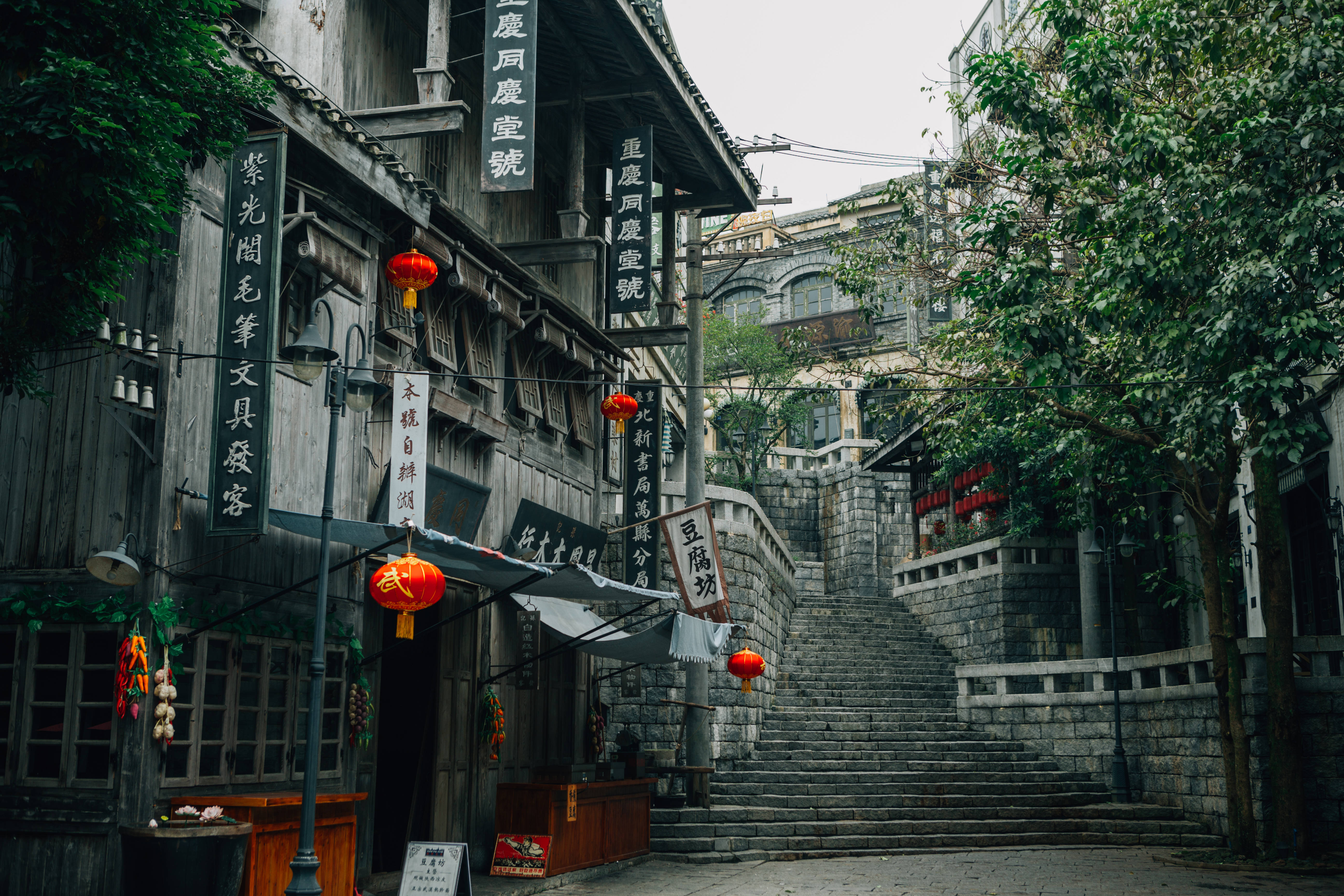 中國 桌布 壁紙 — 一條狹窄的石階梯在風化的木建築之間上升，這些建築裝飾著紅色燈籠和中文招牌，天空多雲。