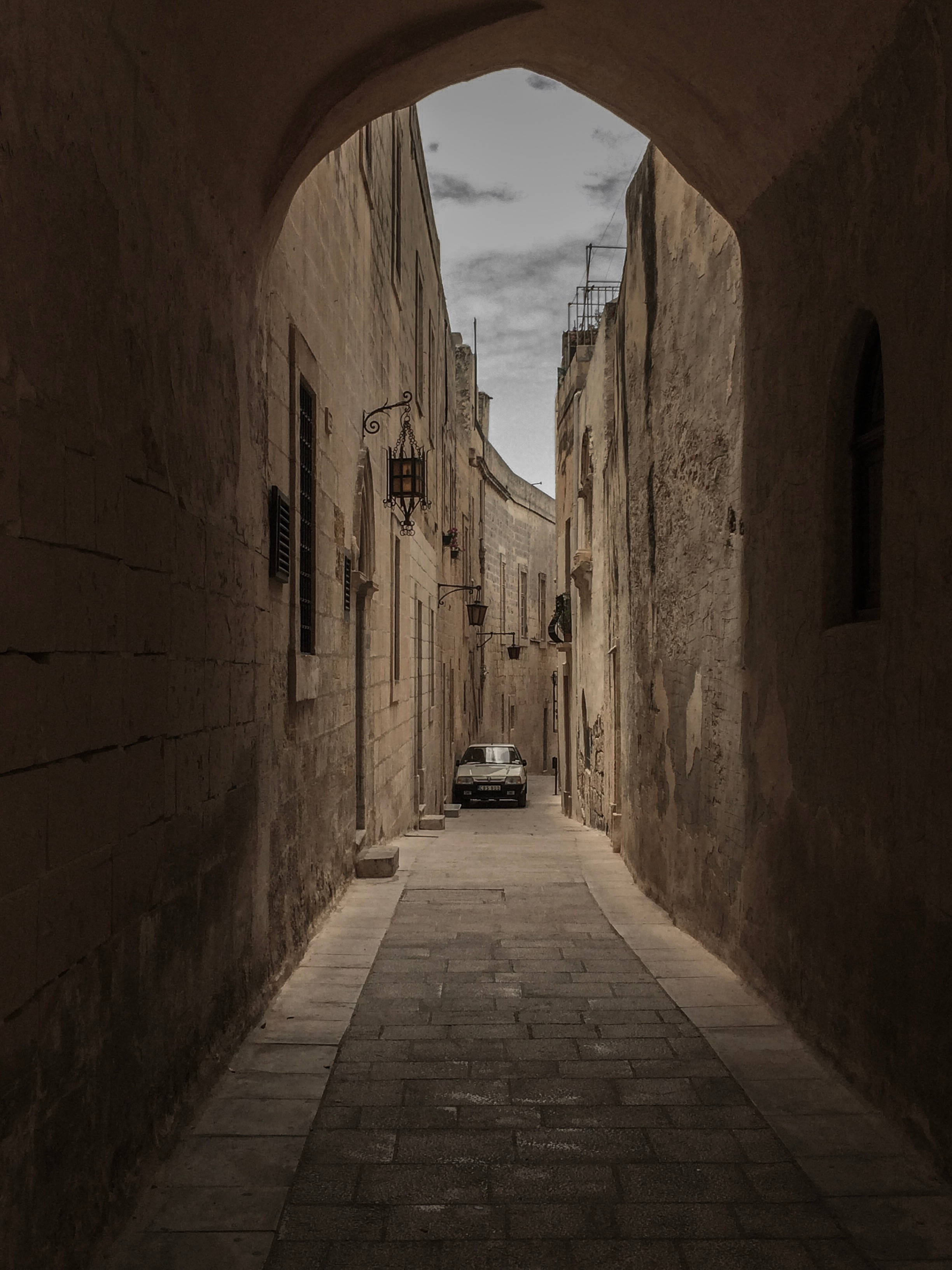 Malta 桌布 壁紙 — 一條狹窄的石頭小巷，由拱形門廊框住，通往一輛停泊在風化建築物之間的經典汽車，天空多雲。