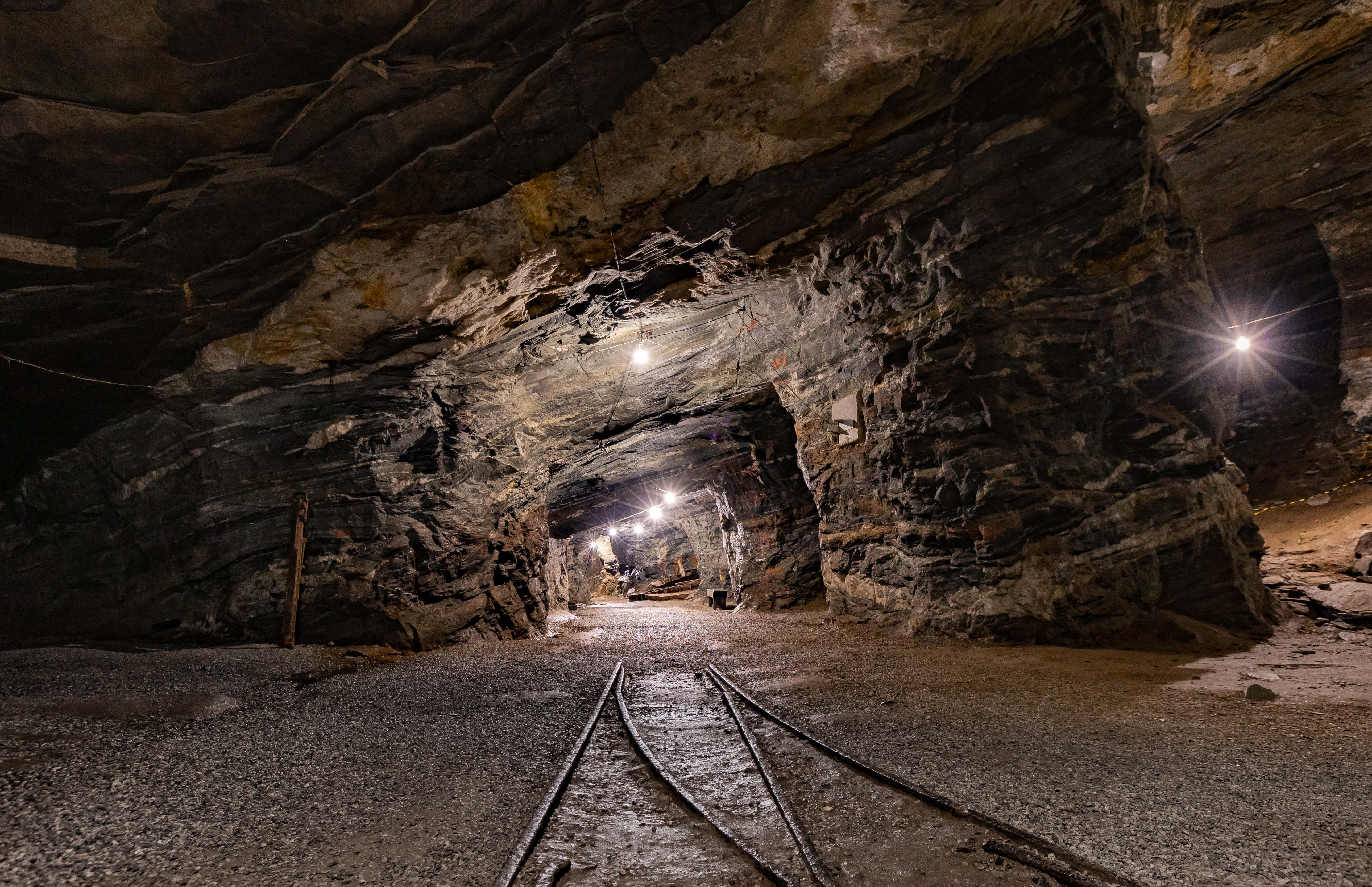 Mining 桌布 壁紙 — 一條狹窄的礦井隧道，粗糙的岩壁和鐵軌在明亮的人造燈光照射下延伸入黑暗。