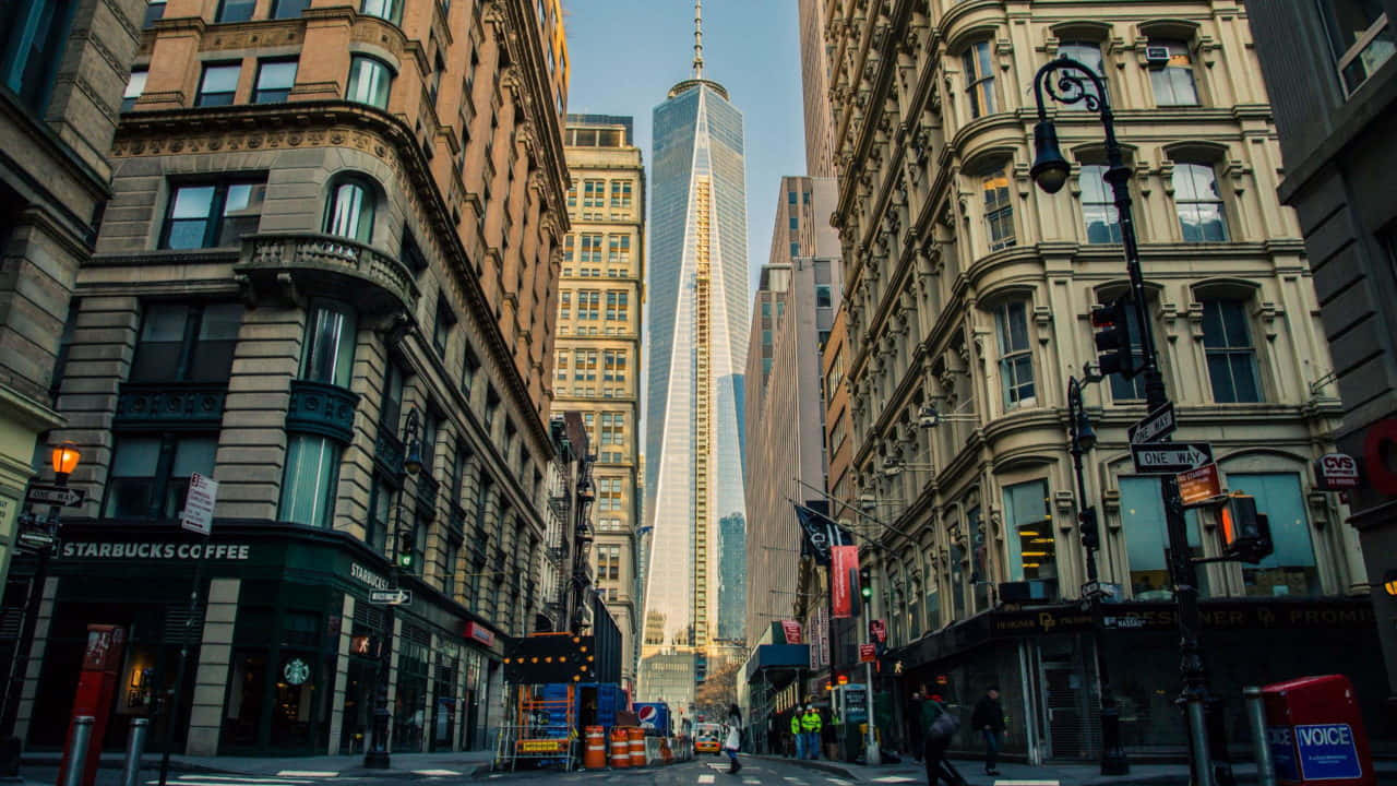720p Ny City 桌布 壁紙 — 一條狹窄的紐約市街道，兩旁是歷史建築，遠處的世貿中心一號大樓在清澈的藍天下高聳入雲。